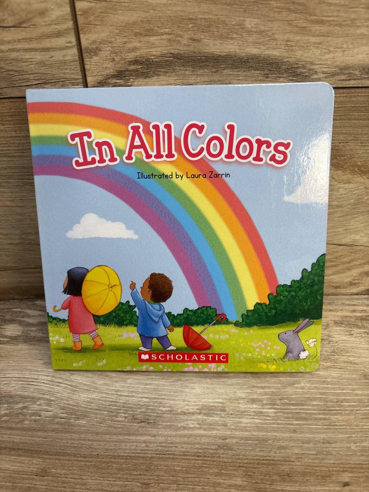Children's board book 'In All Colors' with rainbow, kids, umbrellas, and Scholastic logo