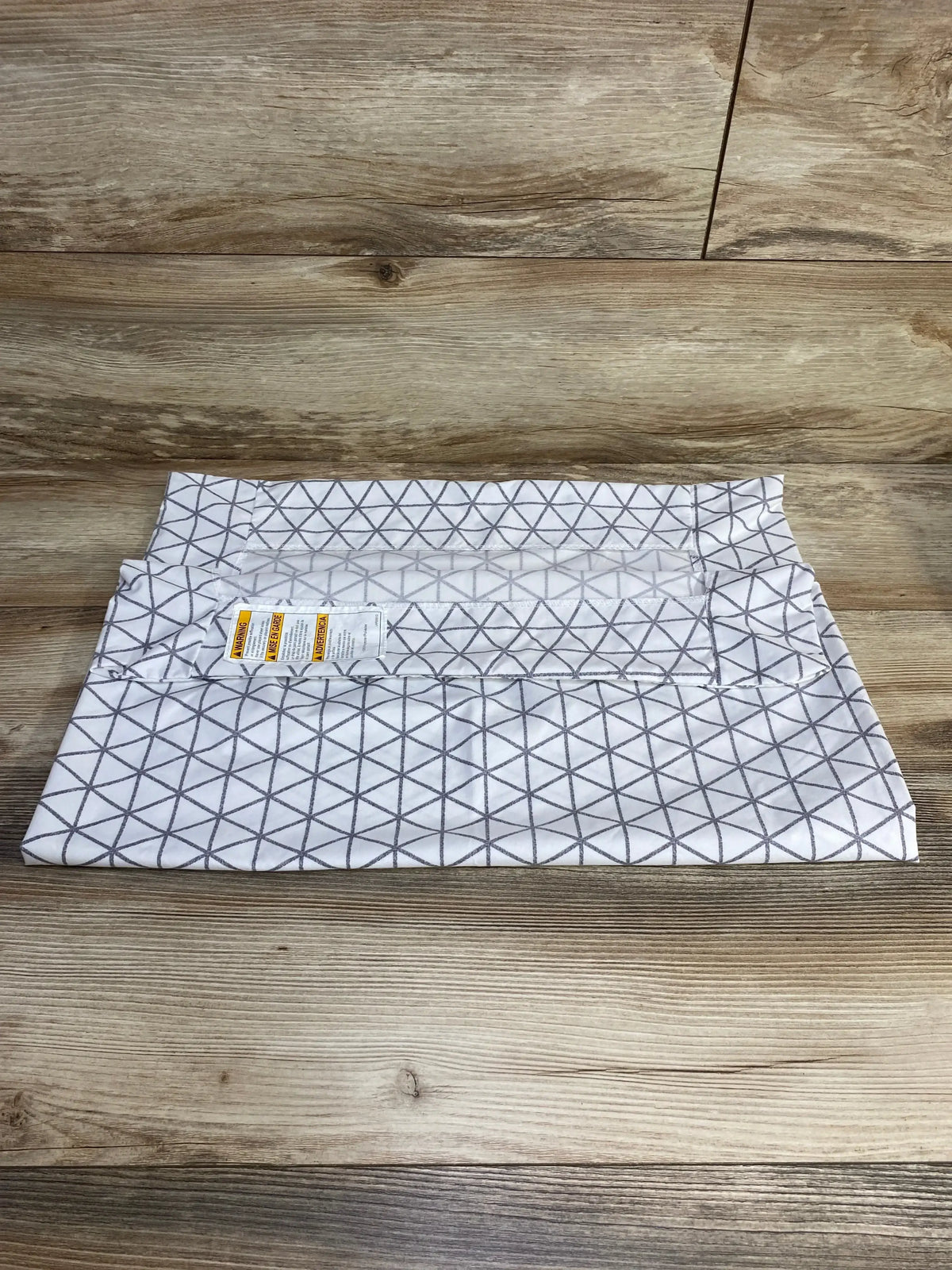 Folded Graco Travel Lite crib sheet, white with grey triangle grid pattern and visible warning label on wood background