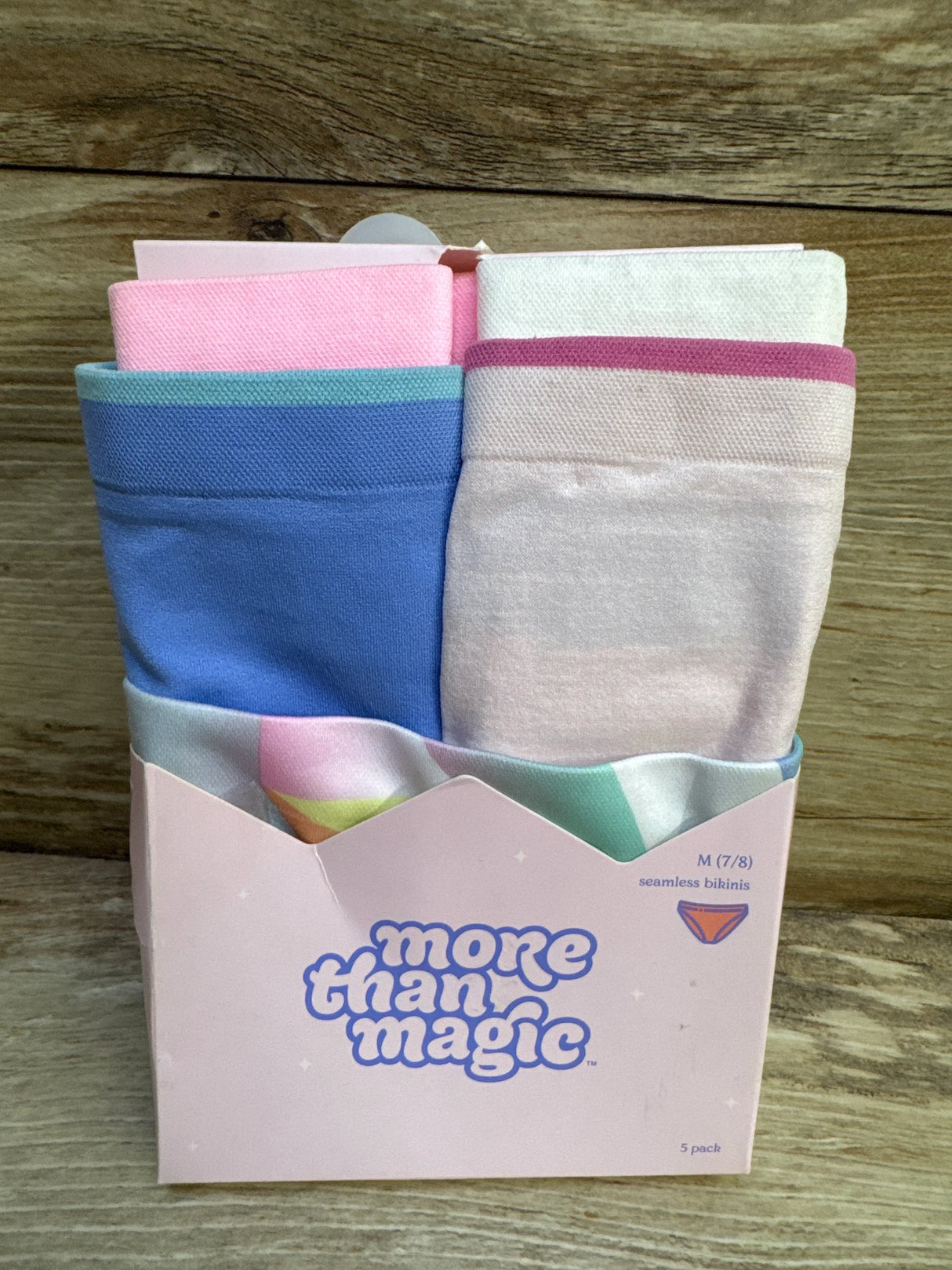 Front view of 5-pack More Than Magic seamless bikinis in pastel blue, pink, and cream inside pink box on wood background