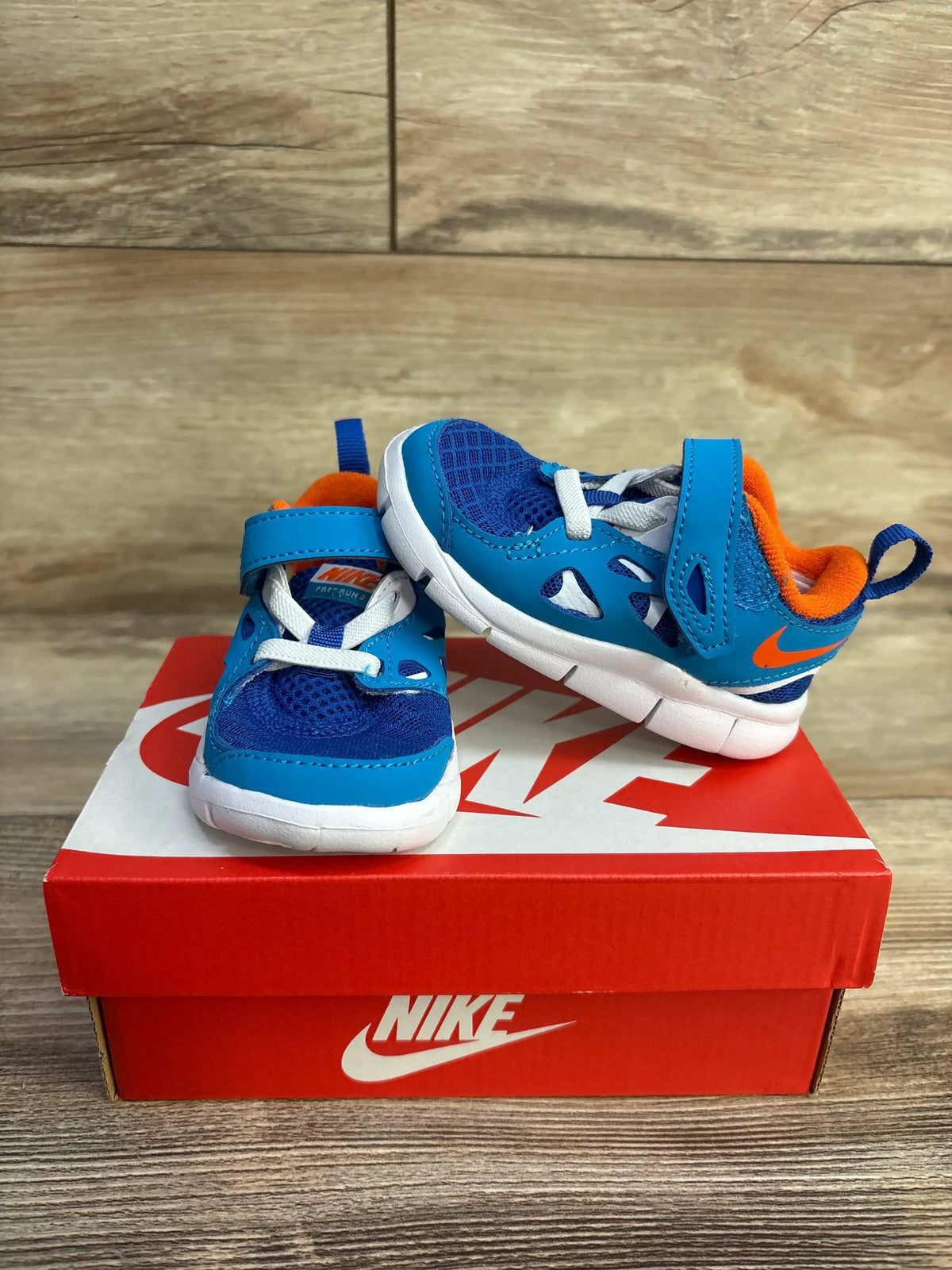 Three-quarter view of Nike Free Run 2 blue and orange toddler sneakers on red Nike shoebox with wood plank background