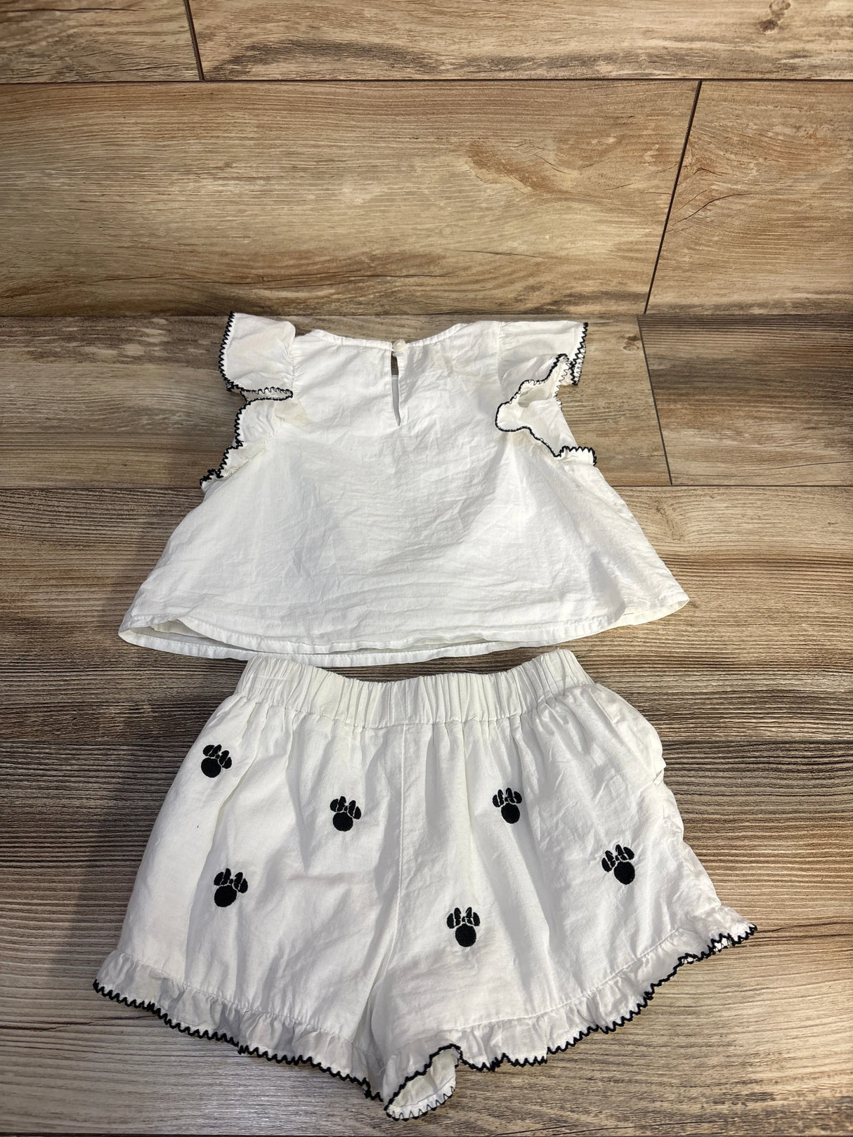 Flat lay of white Minnie Mouse 2-piece top and shorts set showing black embroidered paw icons and black scalloped trim on wood