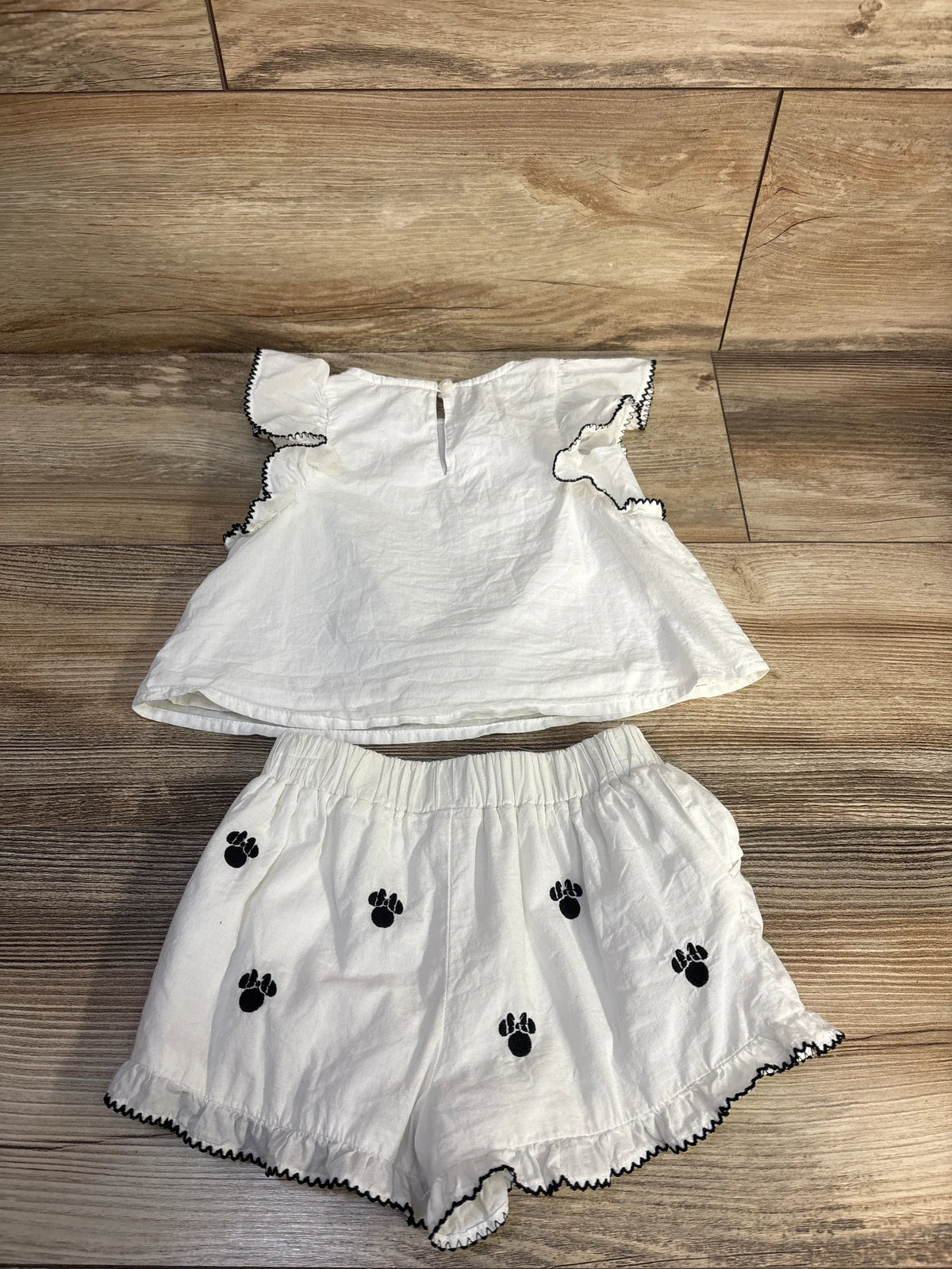 Flat lay of white Minnie Mouse 2-piece top and shorts set showing black embroidered paw icons and black scalloped trim on wood