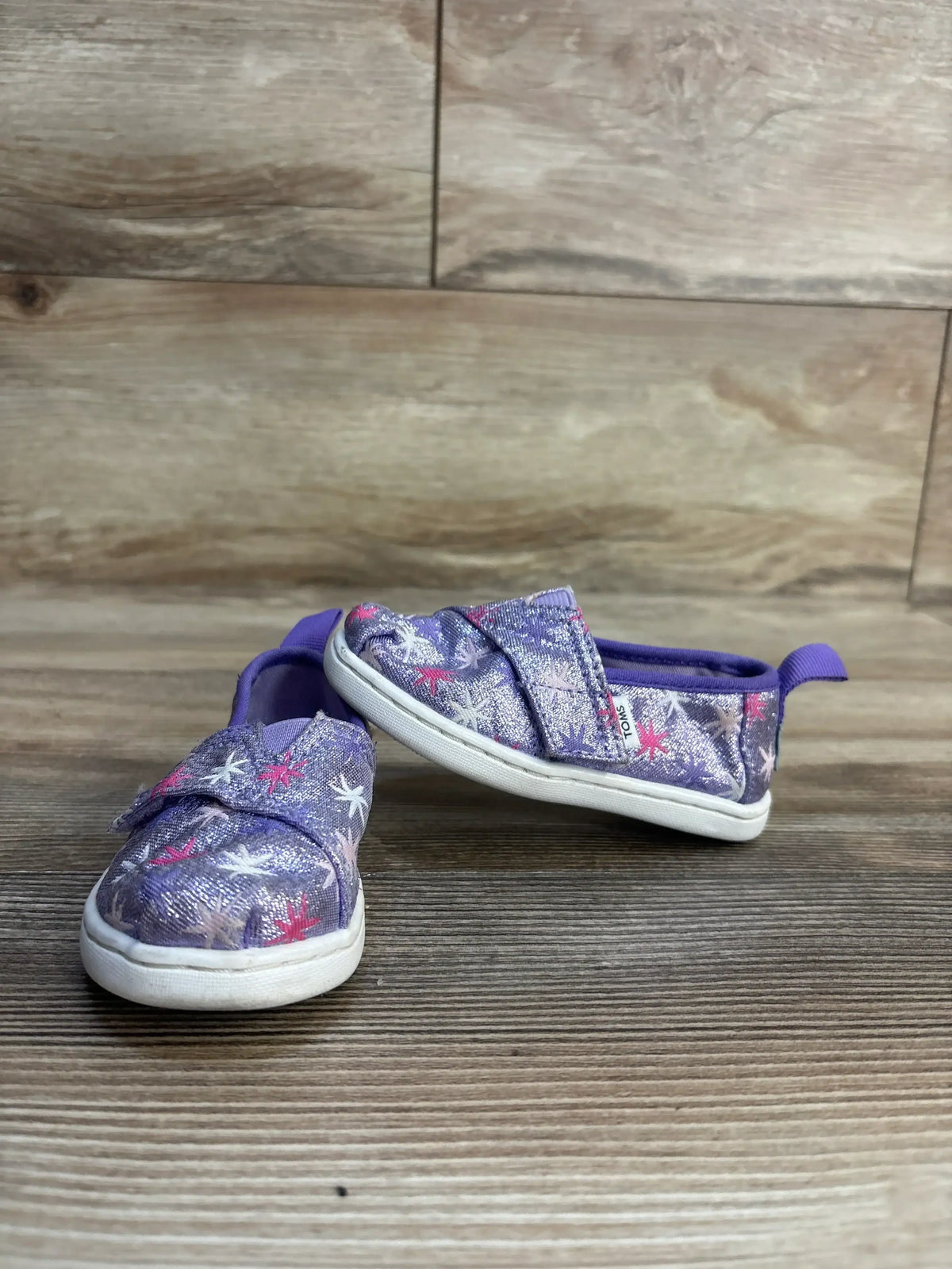 Three-quarter view of Toms Tiny Alpargata toddler shoes, purple glitter with star print and hook-and-loop strap on wood floor