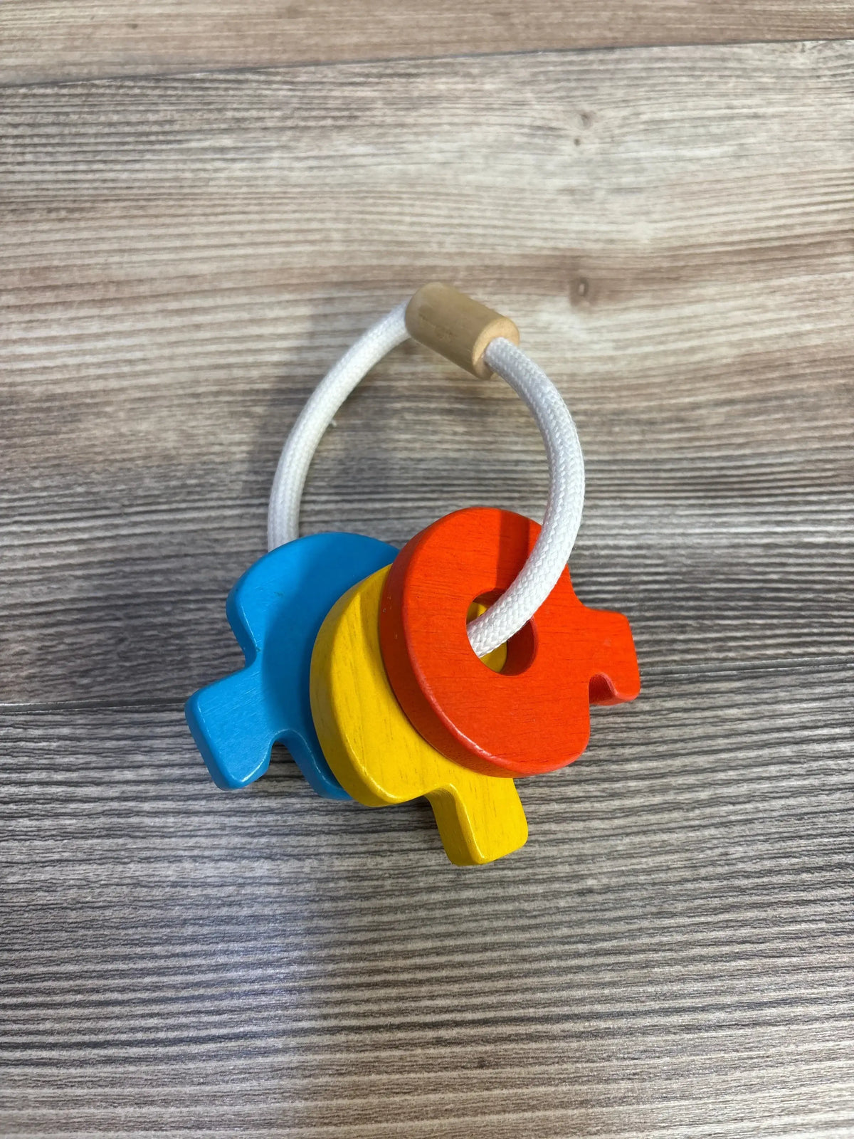 Angled top-down view of wooden baby key rattle with three keys in red, yellow and blue on a white cord over woodgrain surface