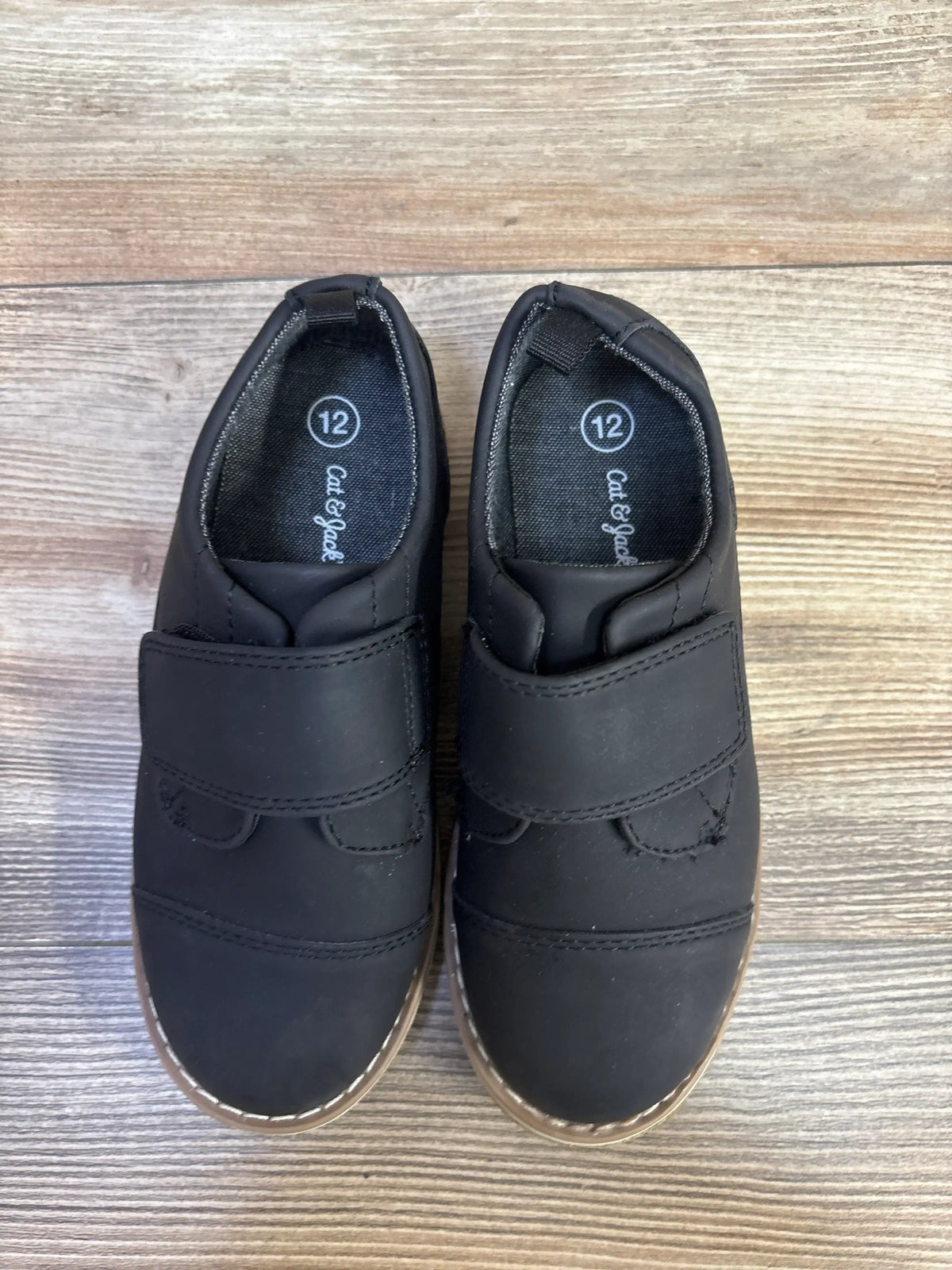 Top-down view of Cat & Jack Adonis toddler derby shoes in black, size 12, showing velcro strap closure and brown stitched sole on wood floor