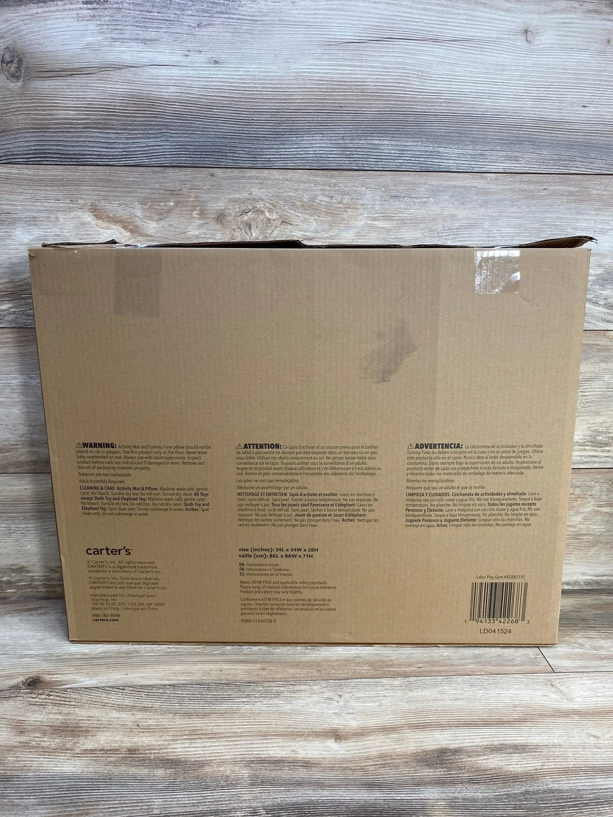 Front view of Carter’s Safari Play Gym box showing printed warnings and barcode, slight dent at top, on wood plank background