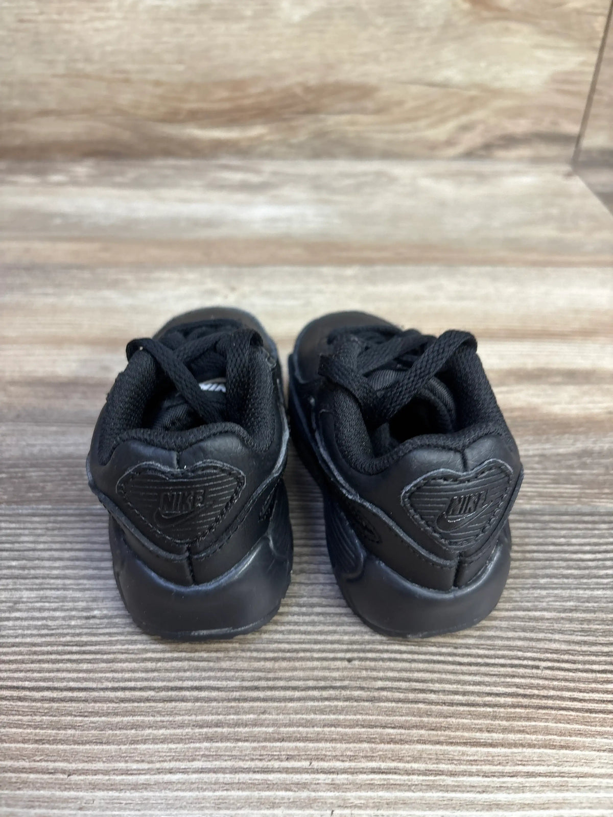 Rear view of Nike Air Max 90 TD sneakers showcasing the Triple Black colorway and rubber outsole on a wood-grain surface