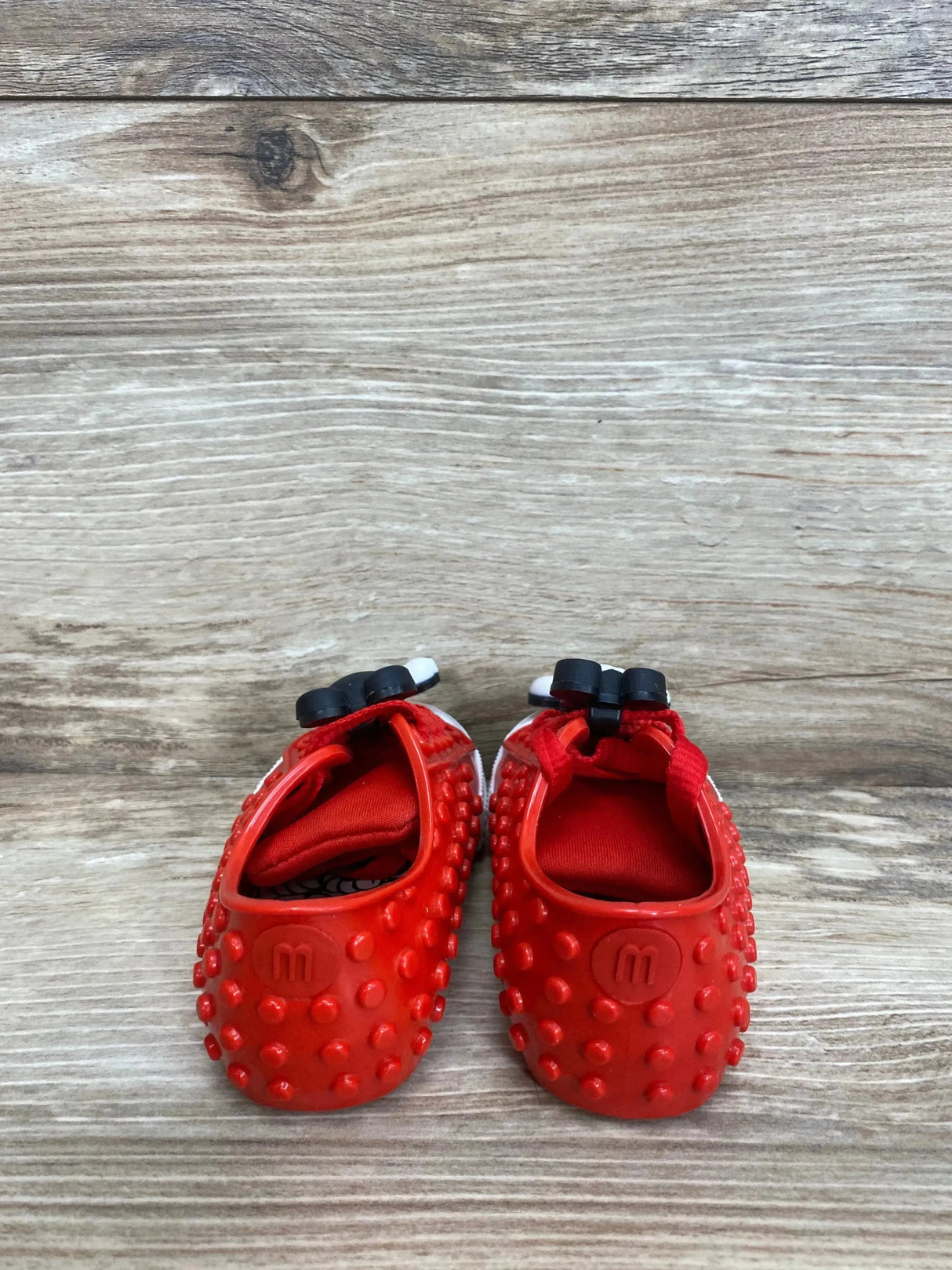Top view of Mini Melissa Mini Polibolha Disney red toddler shoes showcasing raised studded texture and black Minnie bow on wood background