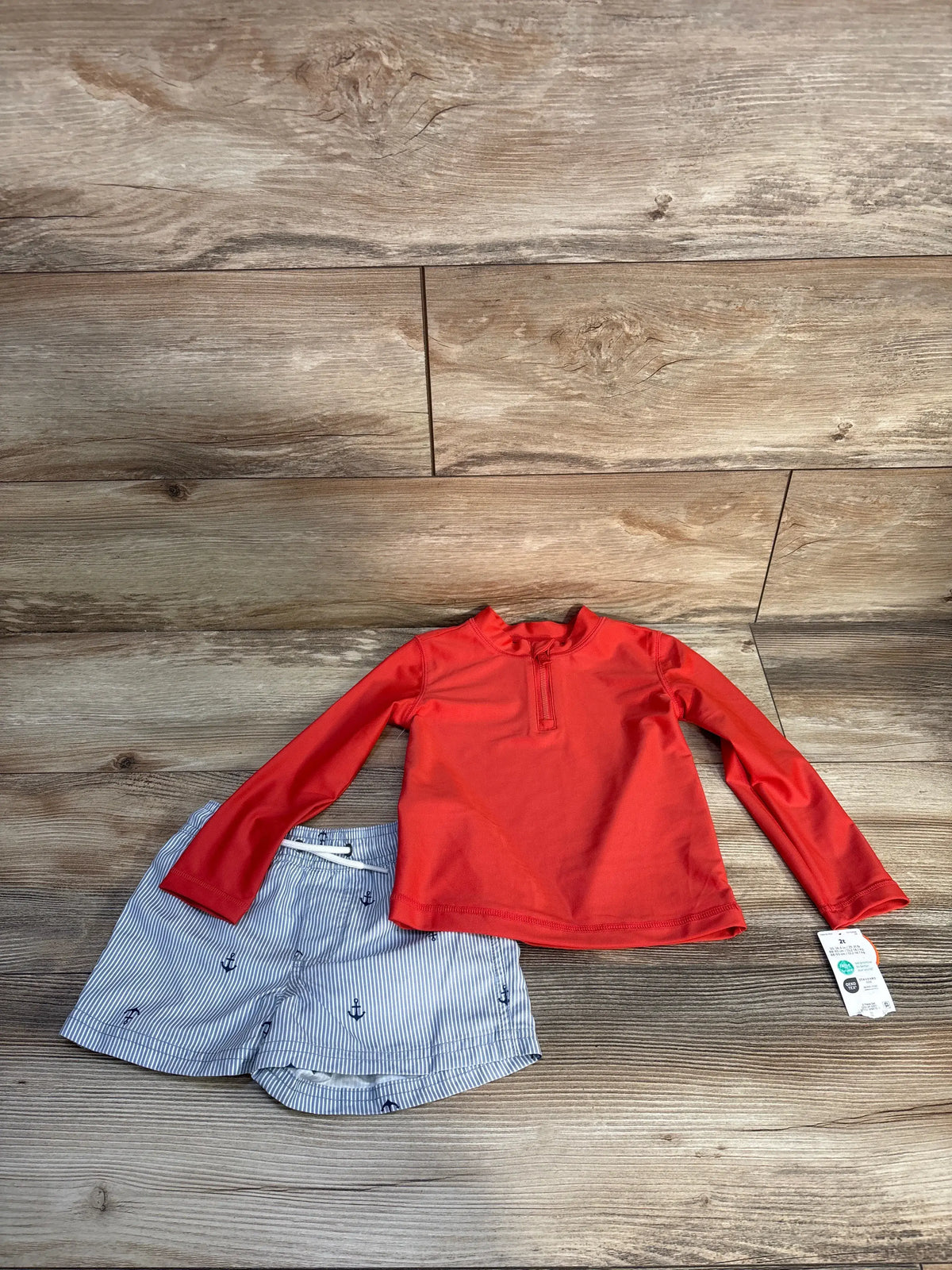 Top-down view of the Just One You rash guard set with red quarter-zip top and striped anchor-print shorts on wood flooring