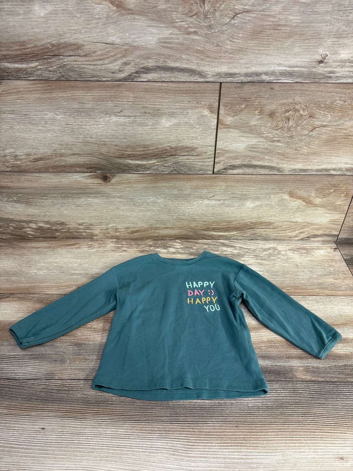 Children's teal long-sleeve shirt with 'HAPPY DAY :) HAPPY YOU' colorful text on front, displayed on wooden floor