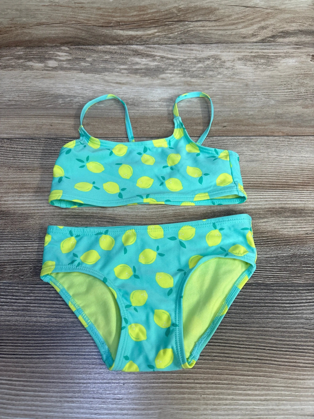 Flat-lay of girls' 2-piece teal lemon-print bikini (2T) showing thin straps and lined full bottoms on wood background