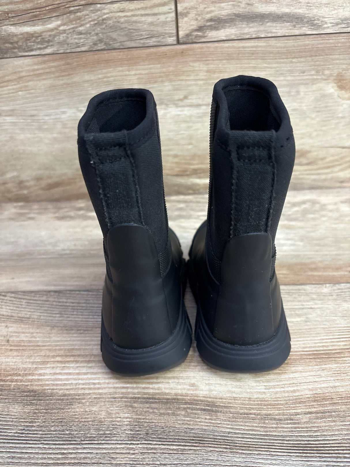 Pair of black children's boots with rubber soles on wood floor, viewed from back