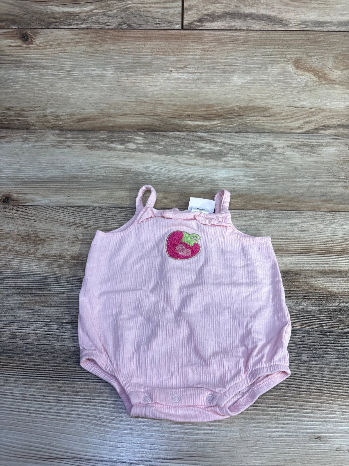 Flat lay of pink Little Lass bubble romper with strawberry patch, ruffle trim, and snap crotch on wood background
