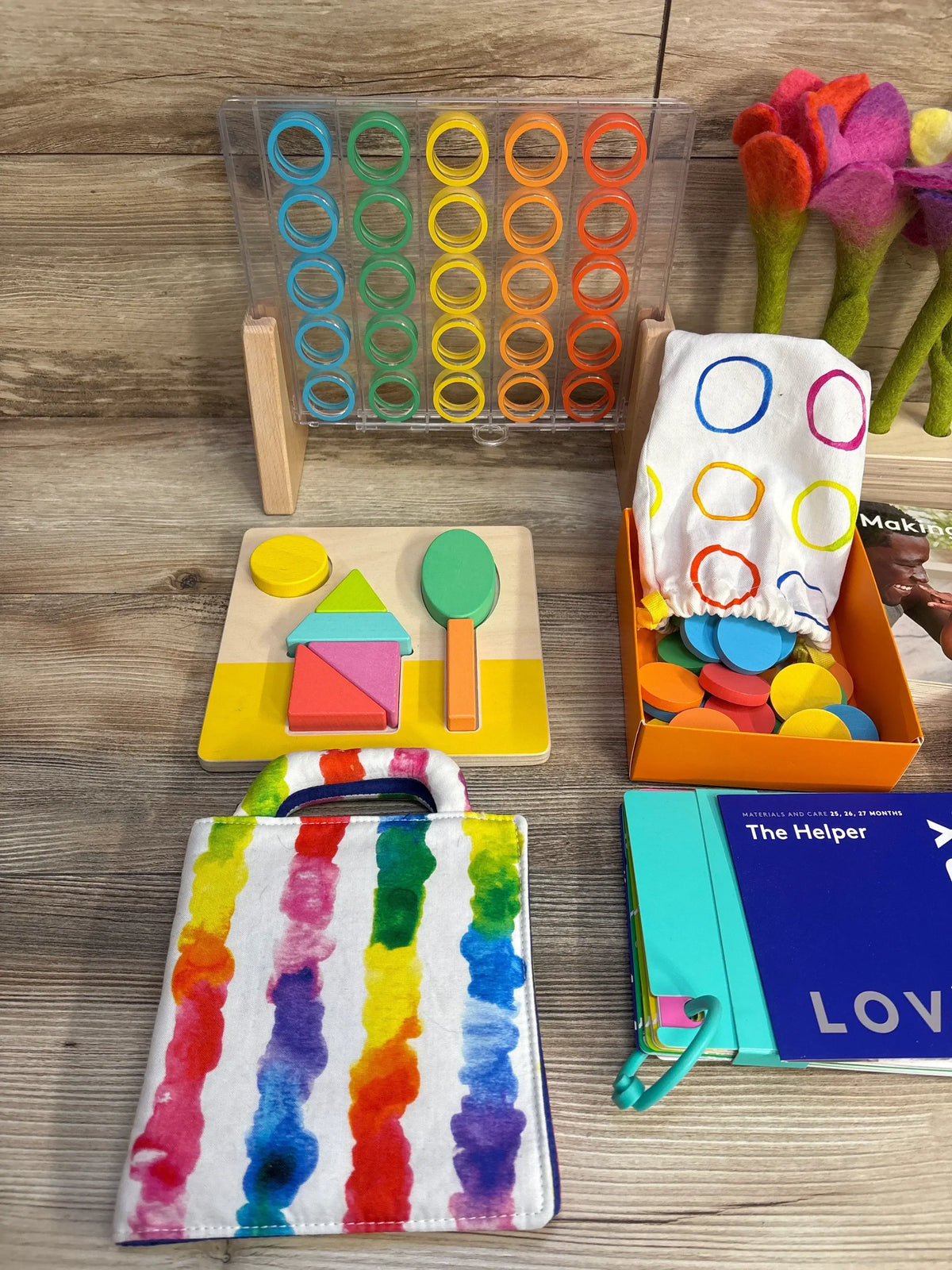 Top view of Lovevery The Helper Play Kit showcasing ring stacker shape puzzle felt flowers rainbow tote and The Helper guidebook on wood plank floor