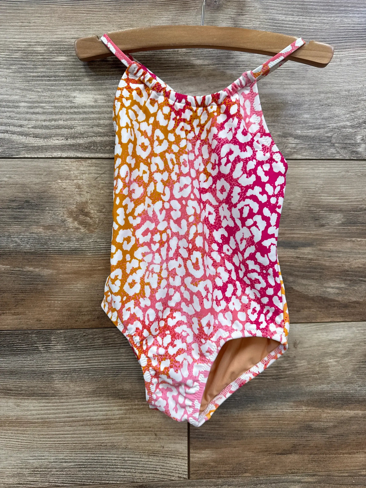 Front view of toddler one-piece swimsuit with orange-to-pink leopard print, thin straps and peach lining on wood backdrop
