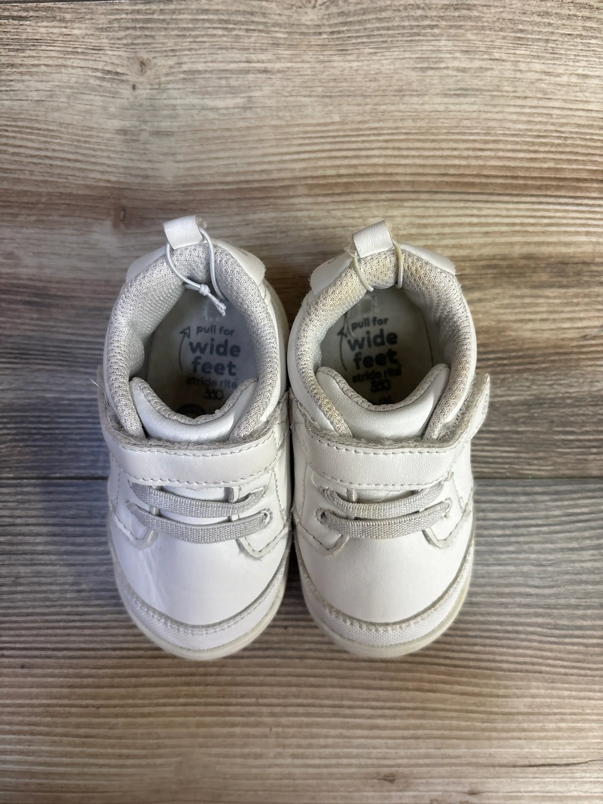 Top view of Stride Rite Taye 2.0 white sneakers with hook-and-loop straps and elastic laces on a wood-grain background