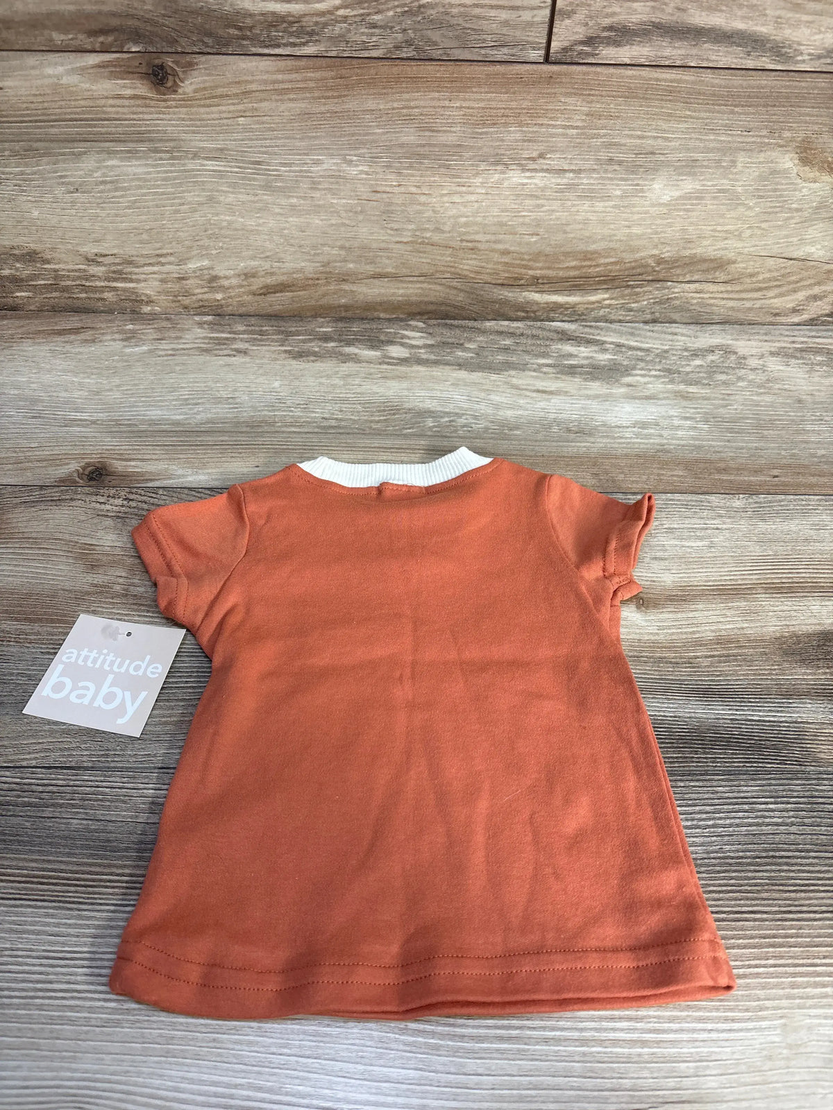 Front view of Attitude Baby short-sleeve shirt in vibrant orange showcasing a white ribbed neckline on a wood-grain background