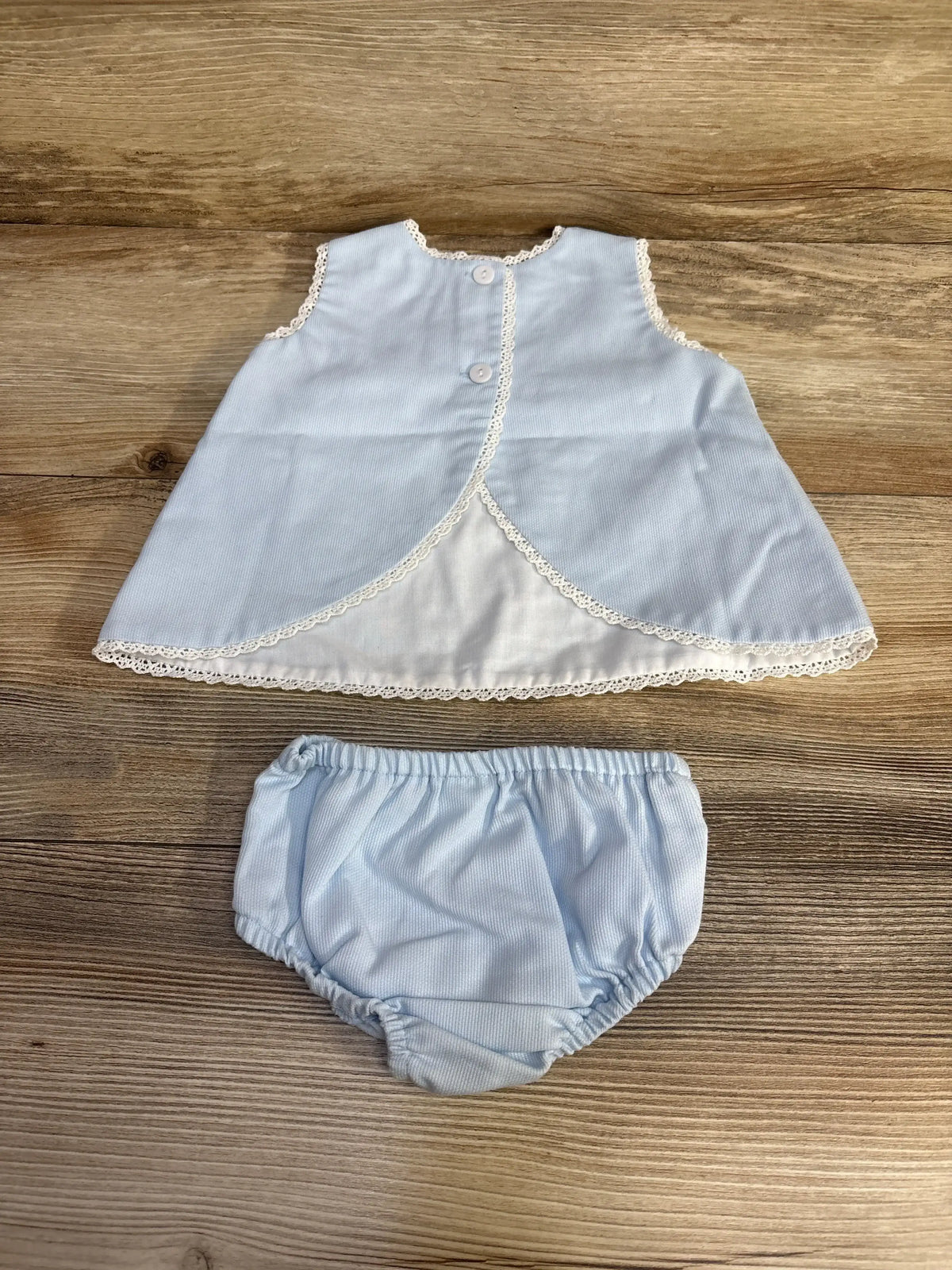 Flat lay of blue Bella Bliss 2pc swing set size 3M, sleeveless top with lace trim and matching bloomers on wood background