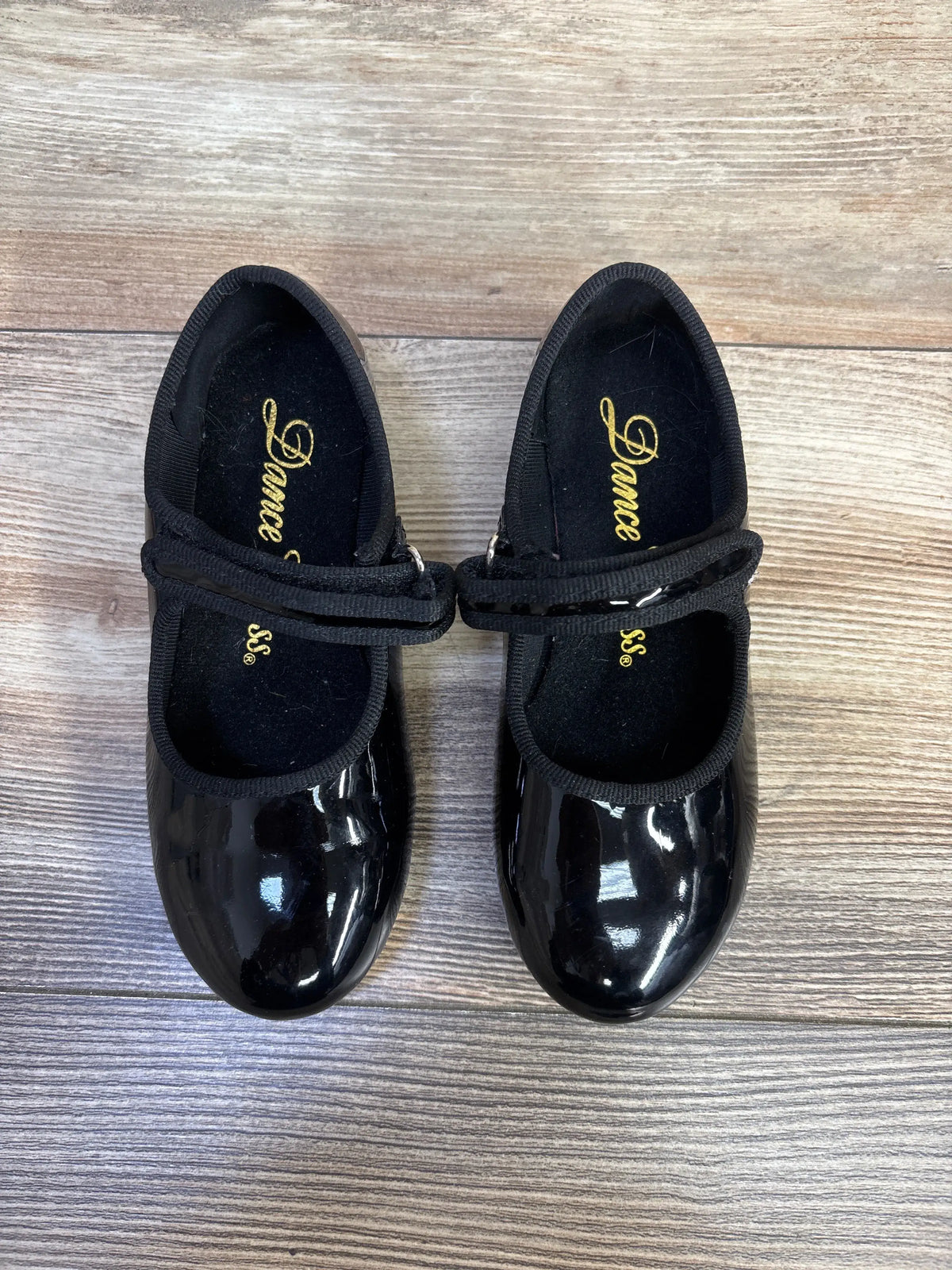 Pair of black patent leather kids' tap dance shoes with strap closures on a wooden background.