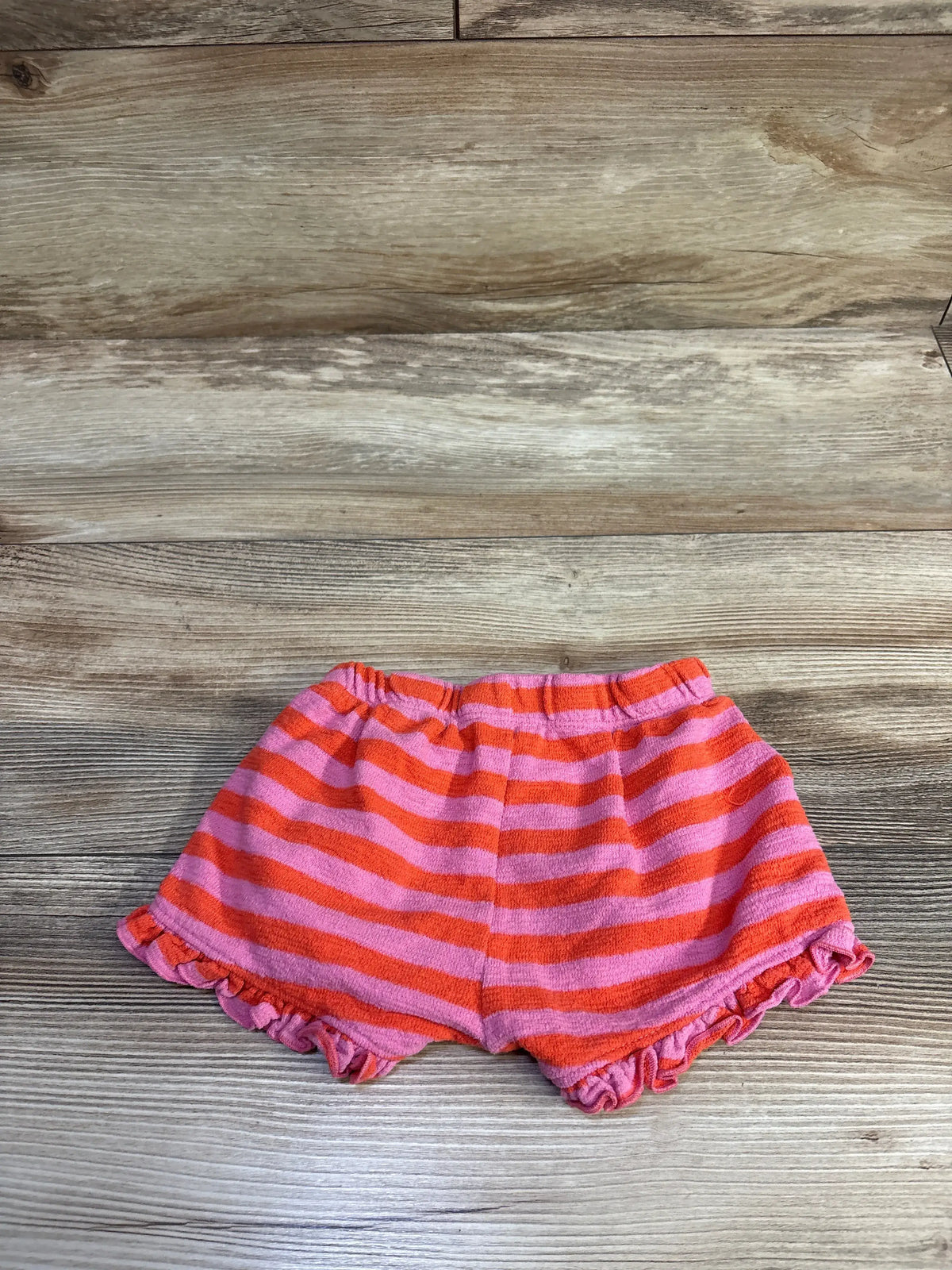 Flat lay of pink and orange striped toddler shorts (18 months) showing elastic waistband and ruffled leg hems on wood background