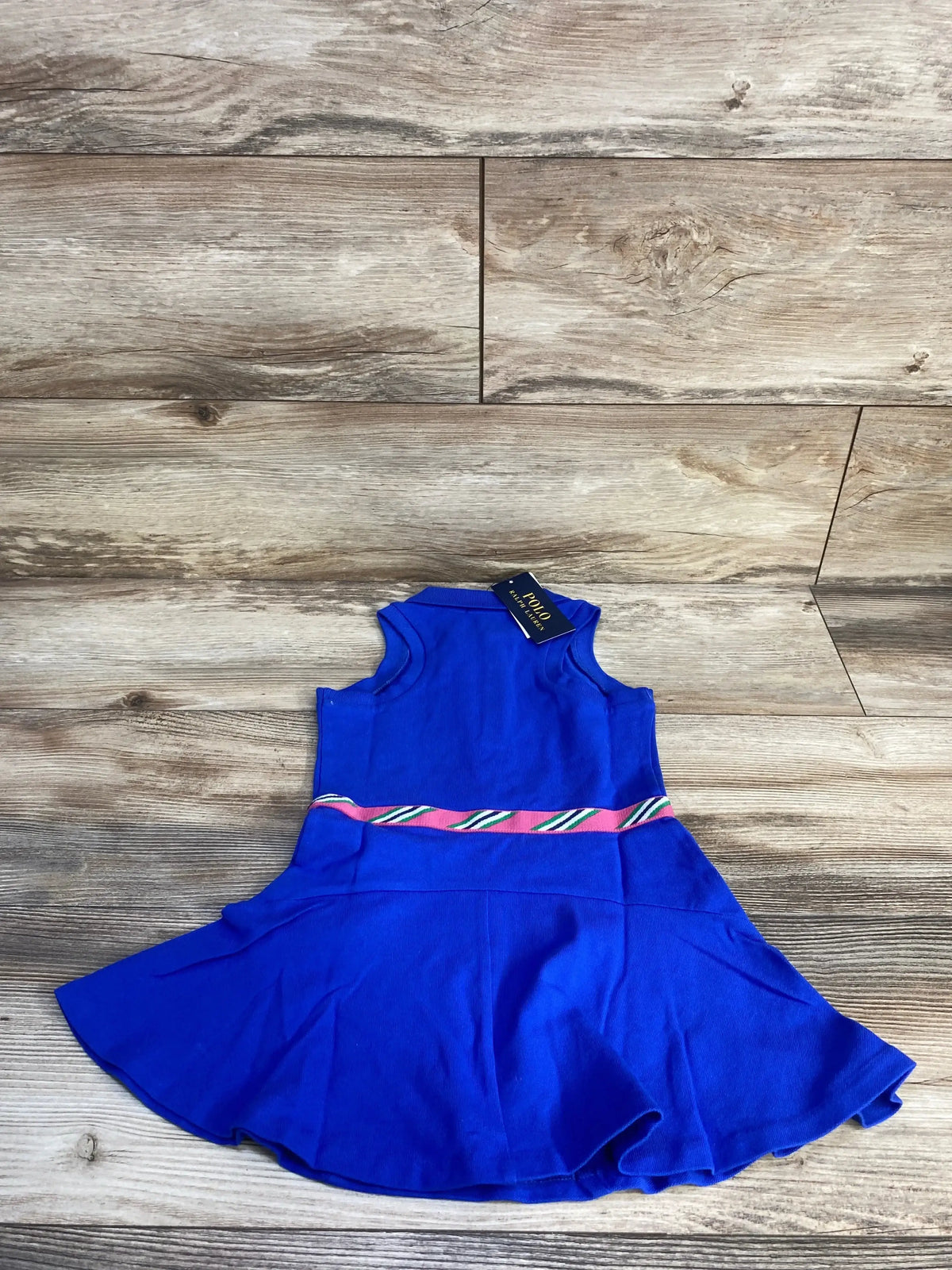 Top-down view of blue Polo Ralph Lauren toddler polo dress with pink-and-green striped waistband, flared skirt on wood floor
