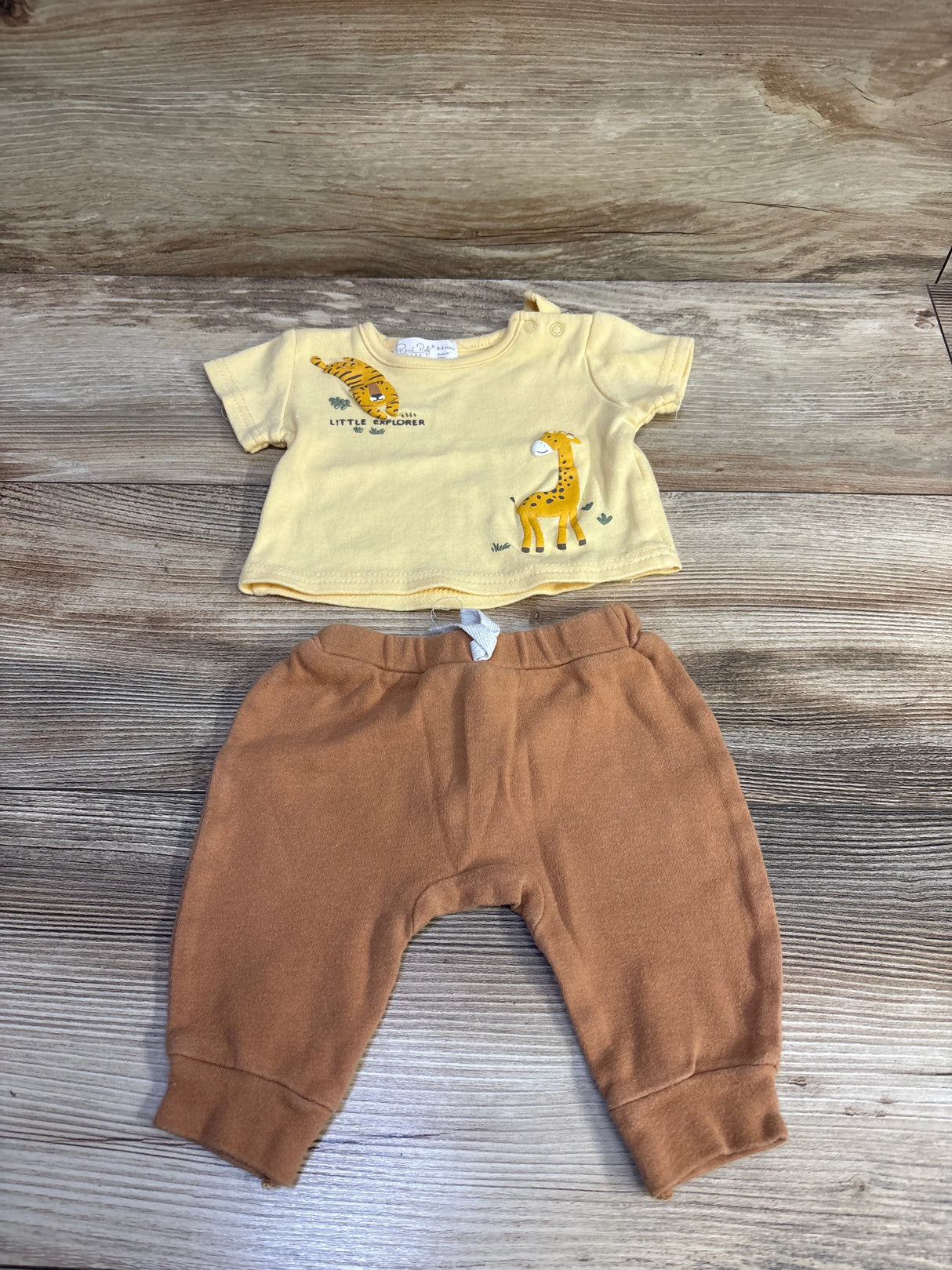 Flat lay of 2-piece baby outfit: yellow Little Explorer shirt with tiger and giraffe prints and brown elastic-waist pants on wood