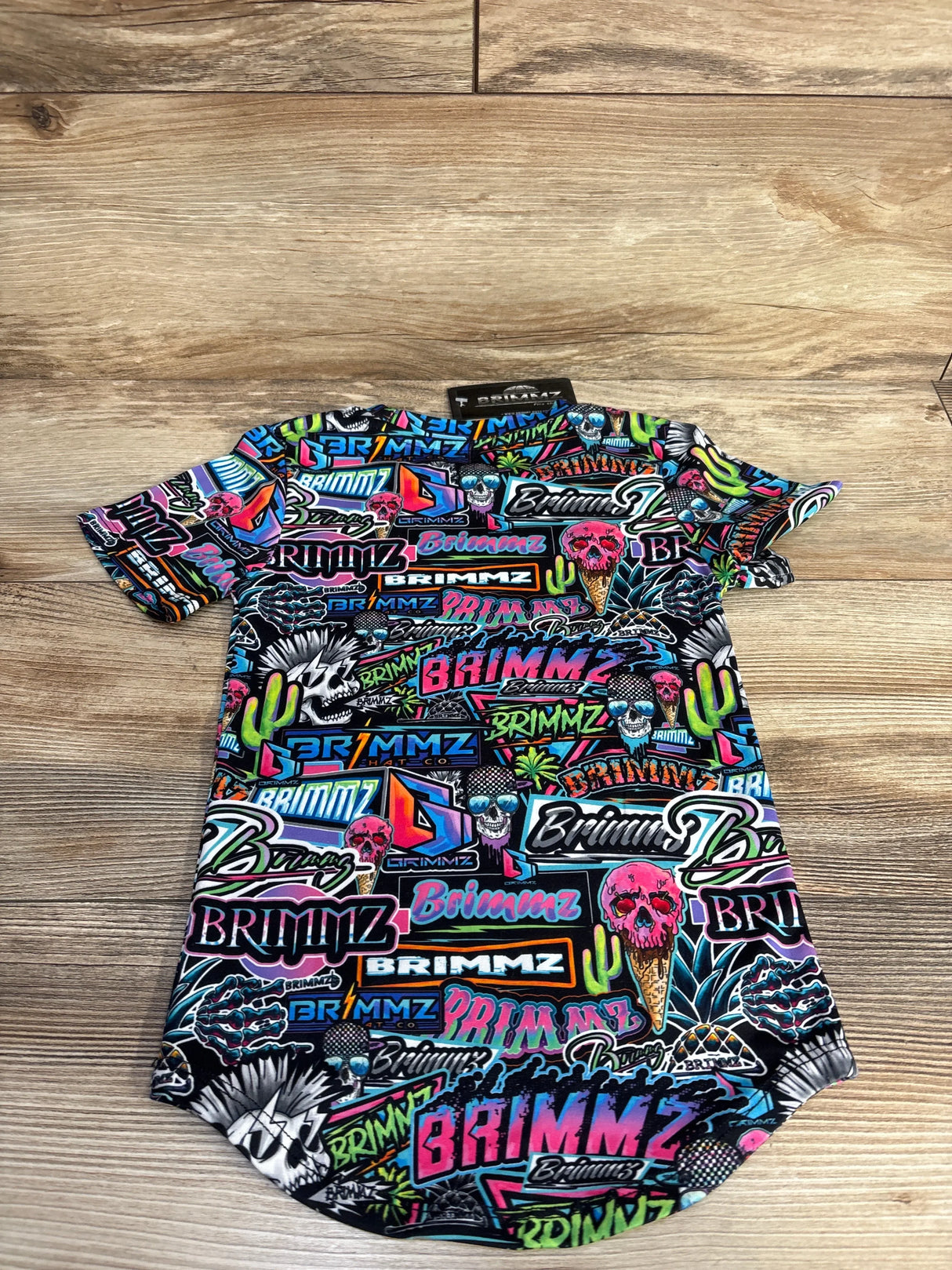 Colorful neon graffiti-style short sleeve shirt with skulls, ice cream cones, cacti, and Brimmz logos on wooden floor
