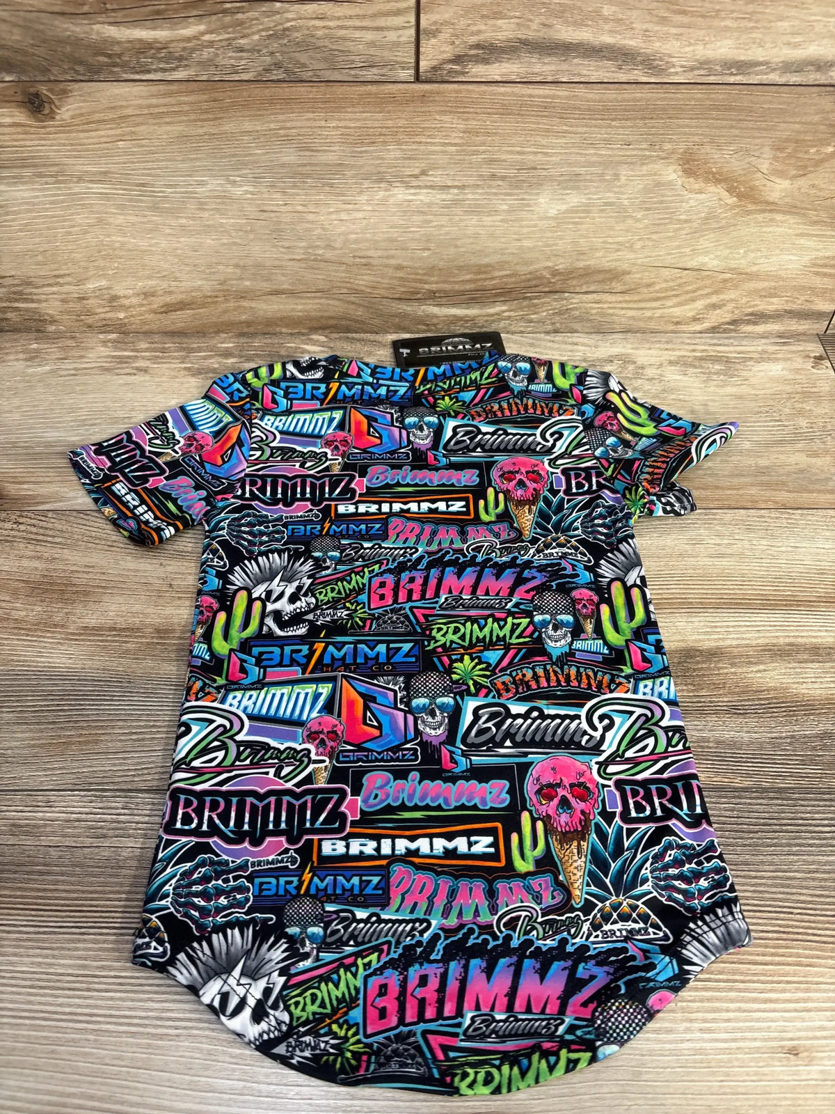 Colorful neon graffiti-style short sleeve shirt with skulls, ice cream cones, cacti, and Brimmz logos on wooden floor