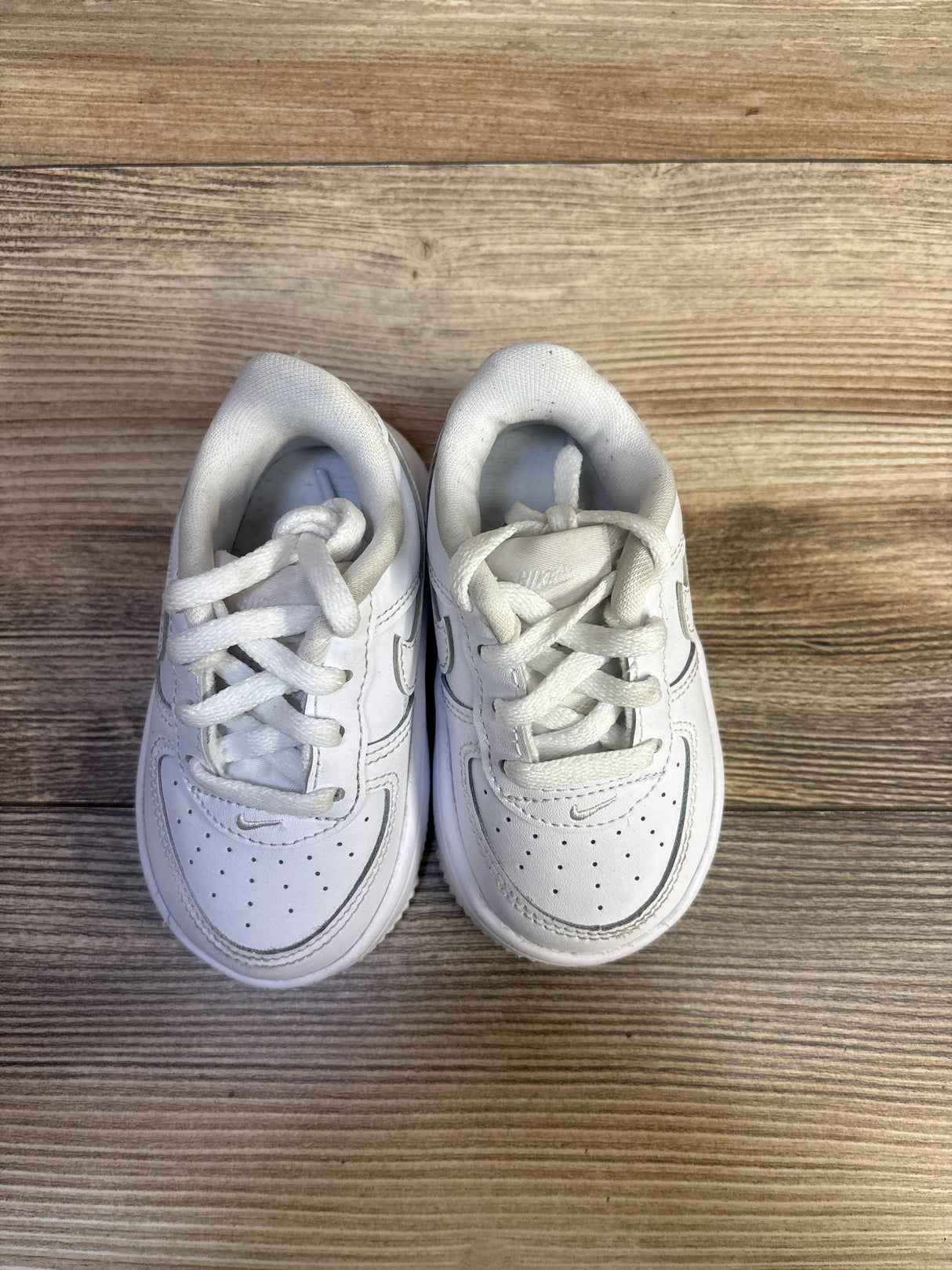 Top-down view of Nike Air Force 1 Low toddler white sneakers showing perforated toe, stitched Swoosh, tied laces on wood floor