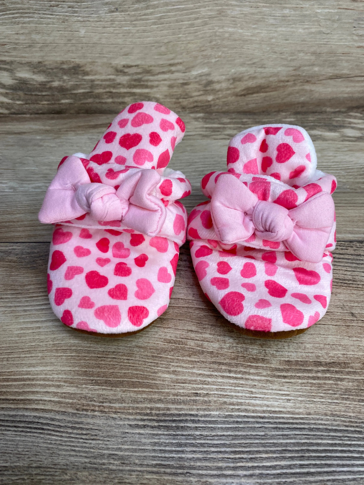Front view of newborn pink cotton booties with heart print and bow trim on wood background, size 6-12 months