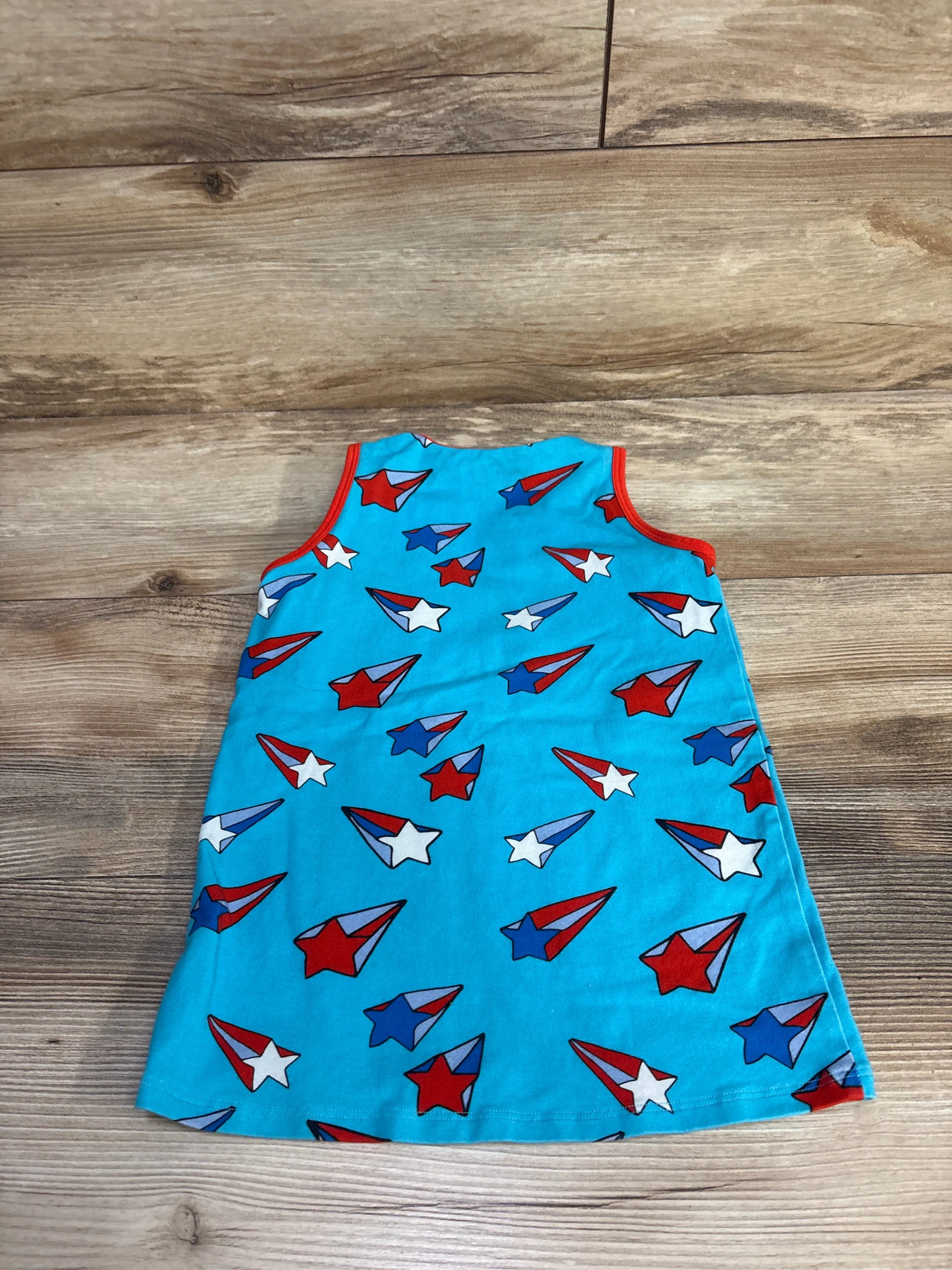 Flat-lay of light blue toddler tank top showing red, white, and blue star and paper-plane print with red trim on wood floor