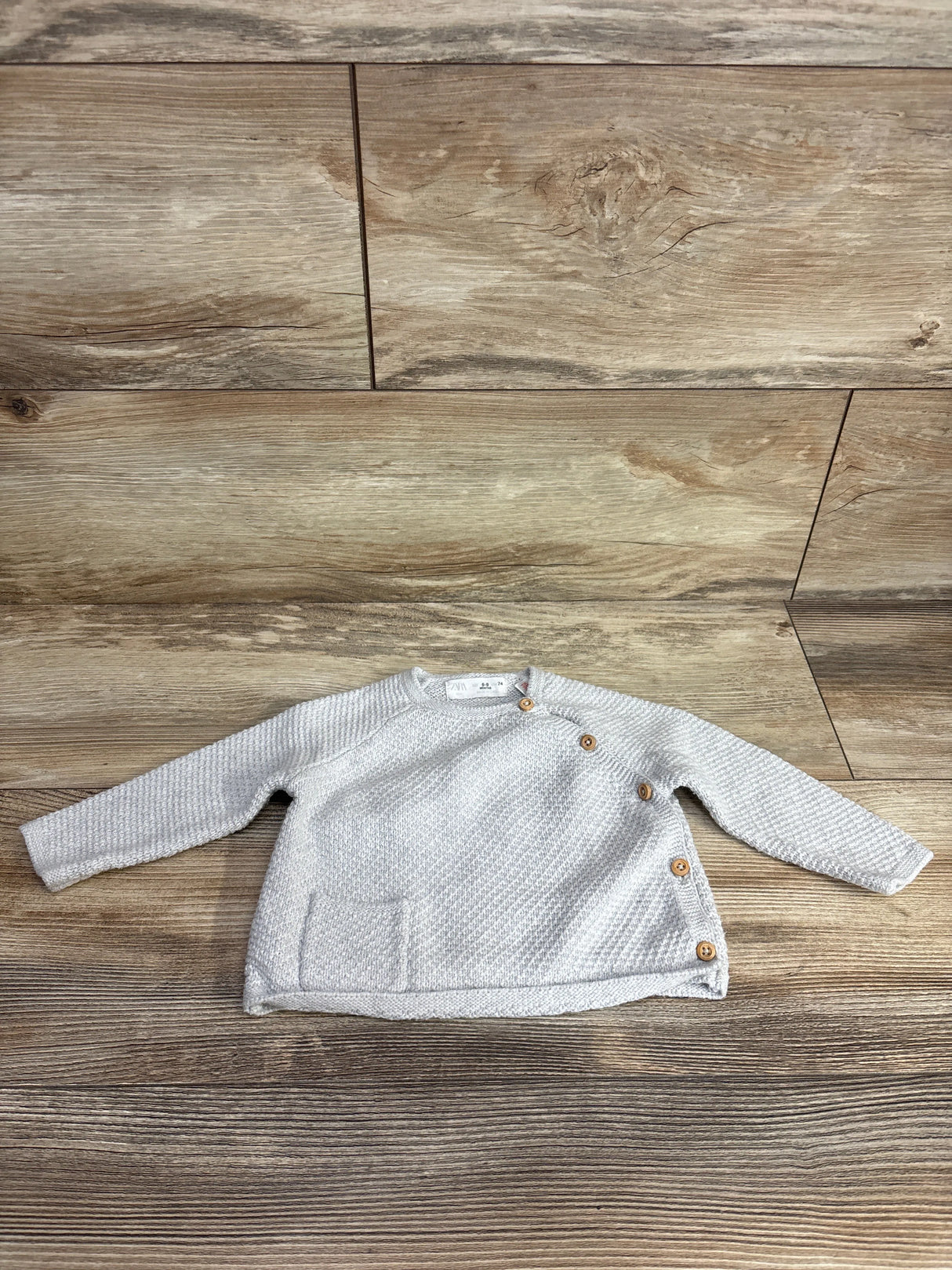 Light gray knit baby sweater with wooden buttons and front pocket on wood background