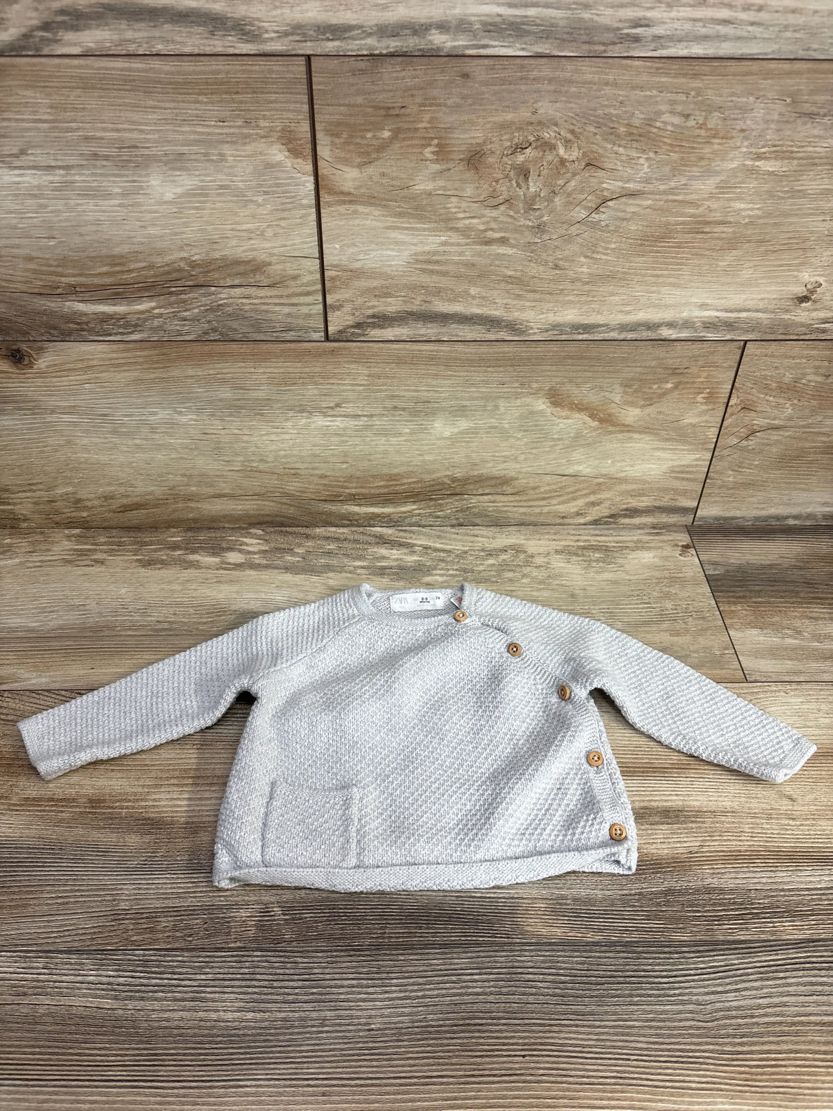 Flat lay of Zara chunky knit kimono baby sweater in light gray showing wooden buttons and front pocket on wood background