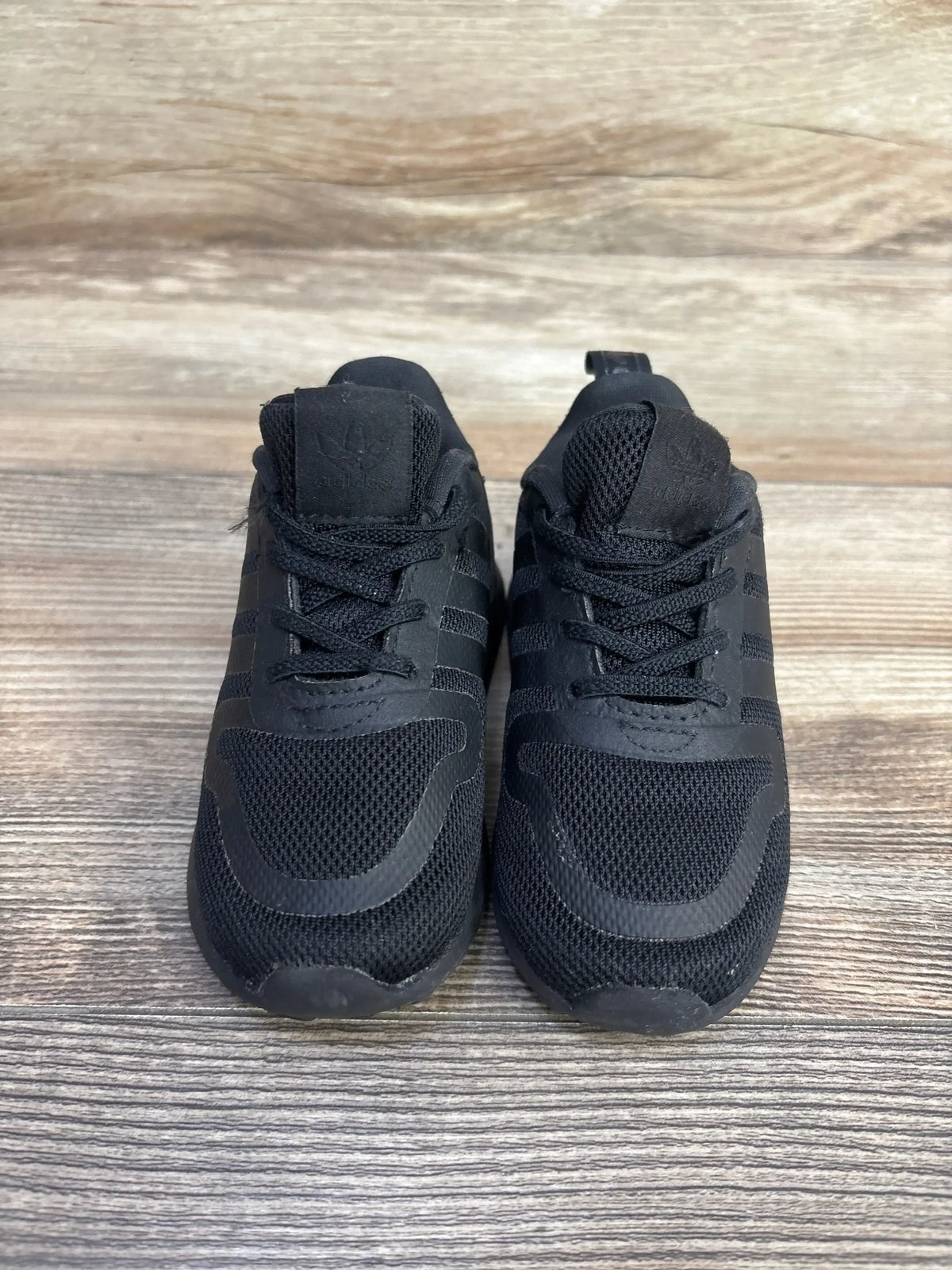 Front view of Adidas Multix I toddler sneakers in triple black showing mesh upper, three stripes, laces on wood background