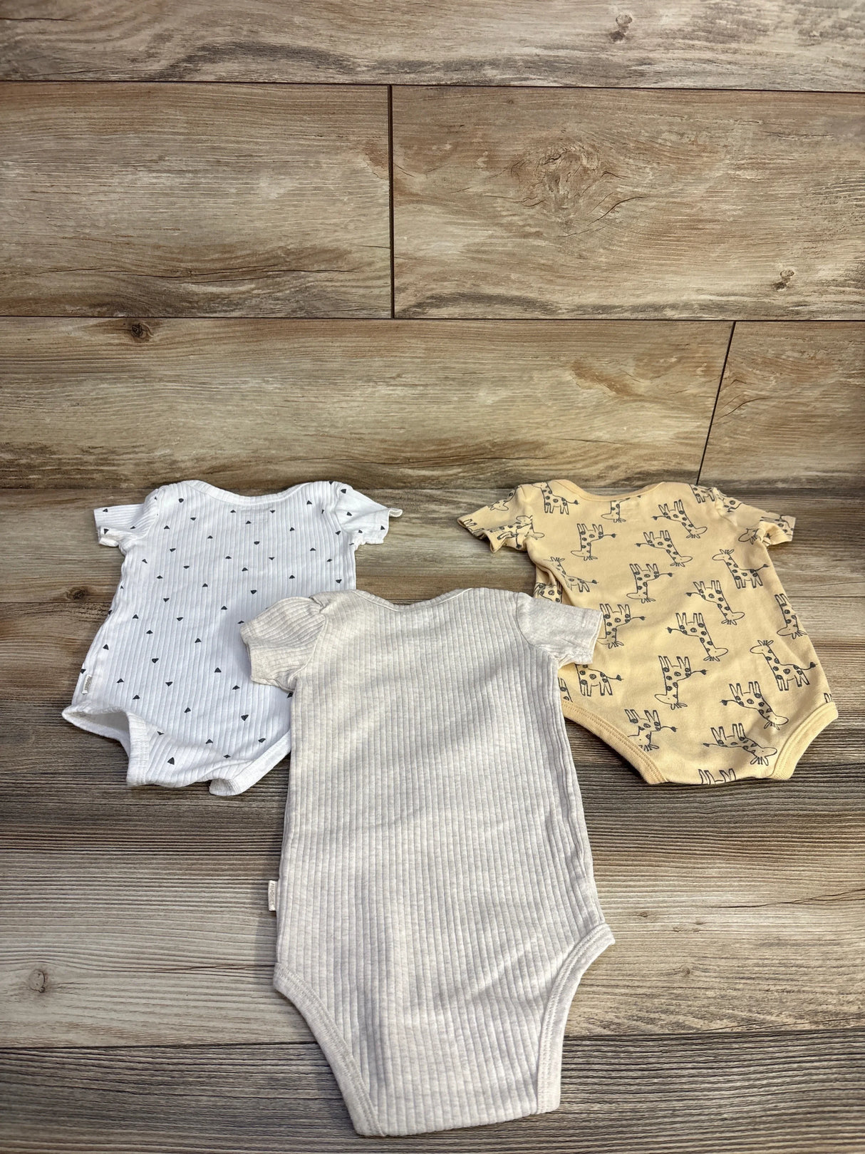Flat lay of Huggies 3-pack bodysuits size 12m showing giraffe print, white hearts, and ribbed beige on wood background