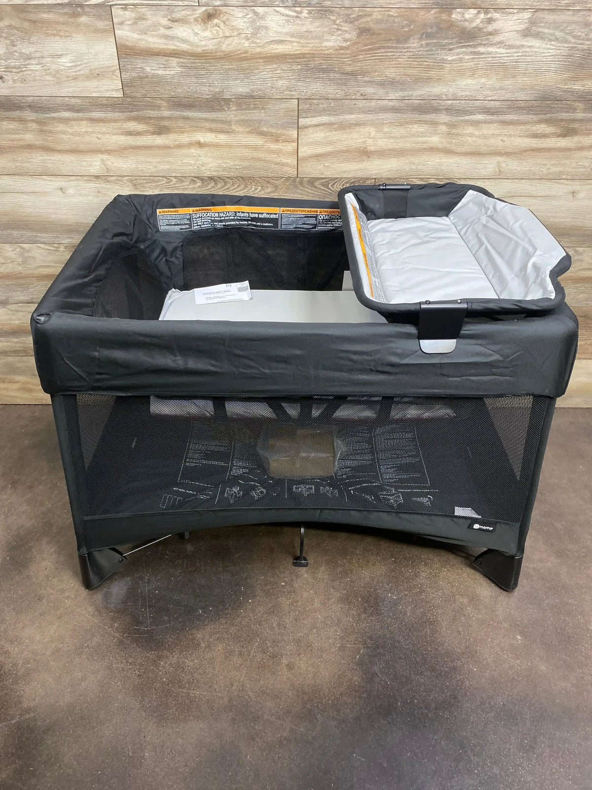 Front view of 4moms Breeze Plus playard in black showing mesh sides and attached changing tray against wood plank wall