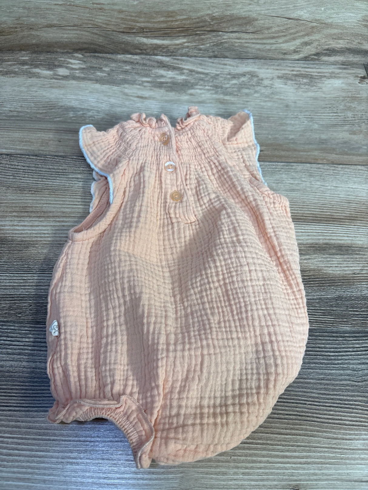 Top down view of peach smocked bubble romper showcasing ruffled sleeves, back buttons and elasticized leg openings on wood surface