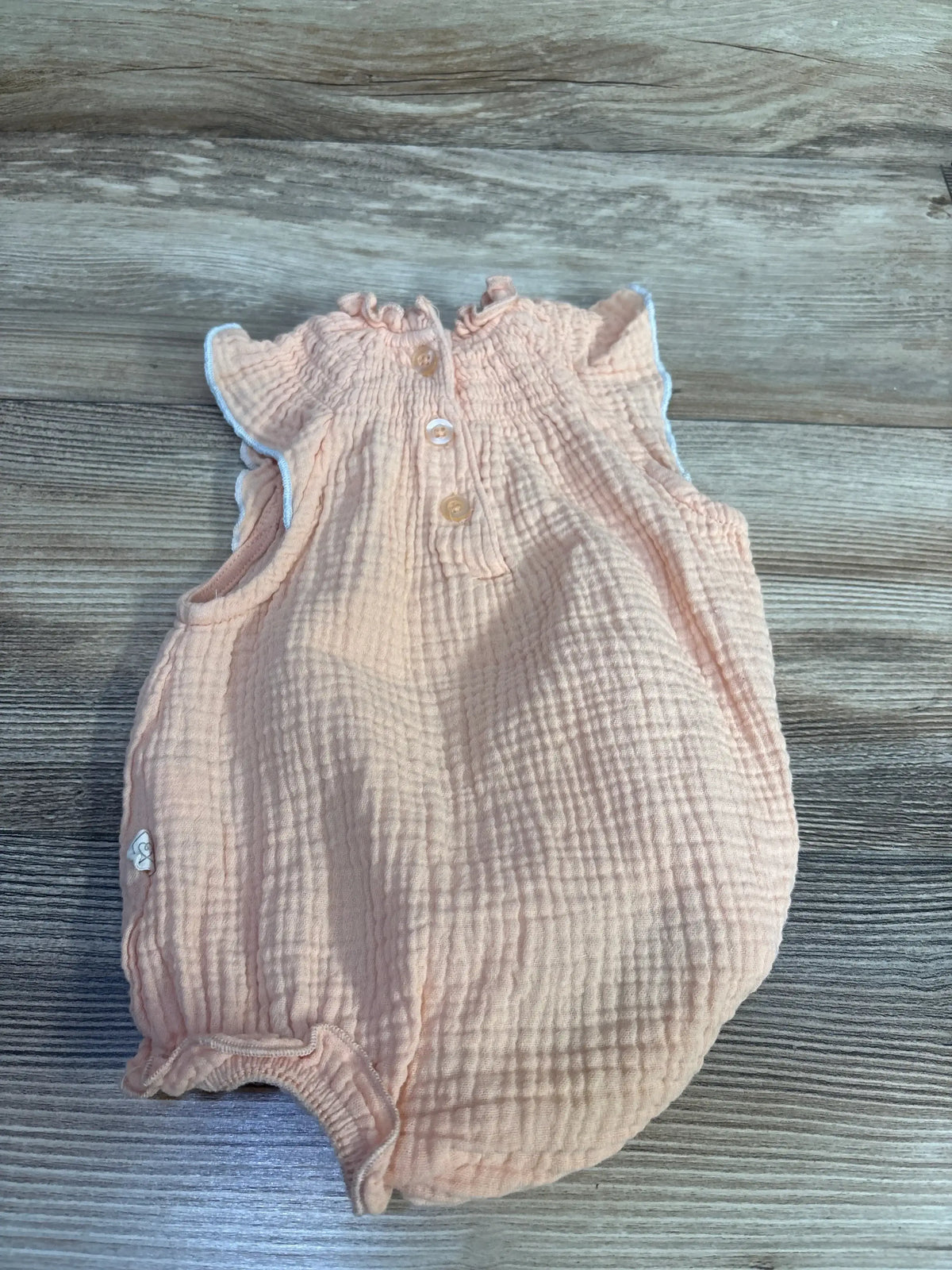 Top down view of peach smocked bubble romper showcasing ruffled sleeves, back buttons and elasticized leg openings on wood surface