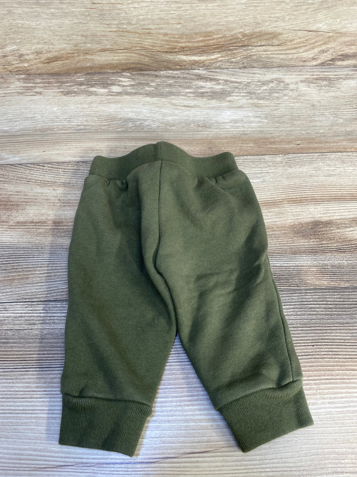 Flat lay of Baby Gap olive drawstring joggers 3 to 6 months showing elastic waistband and ribbed ankle cuffs on wood background