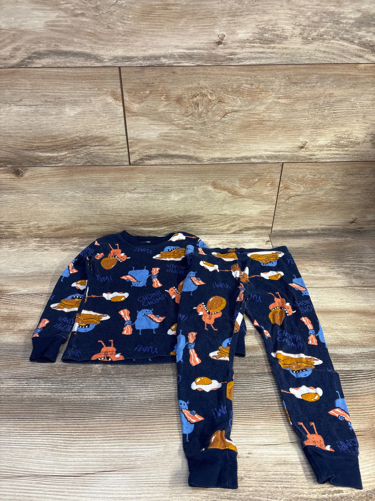 Flat lay of Carter’s navy 2-piece toddler pajamas (3T) showing bacon, egg, and pancake print on wood floor