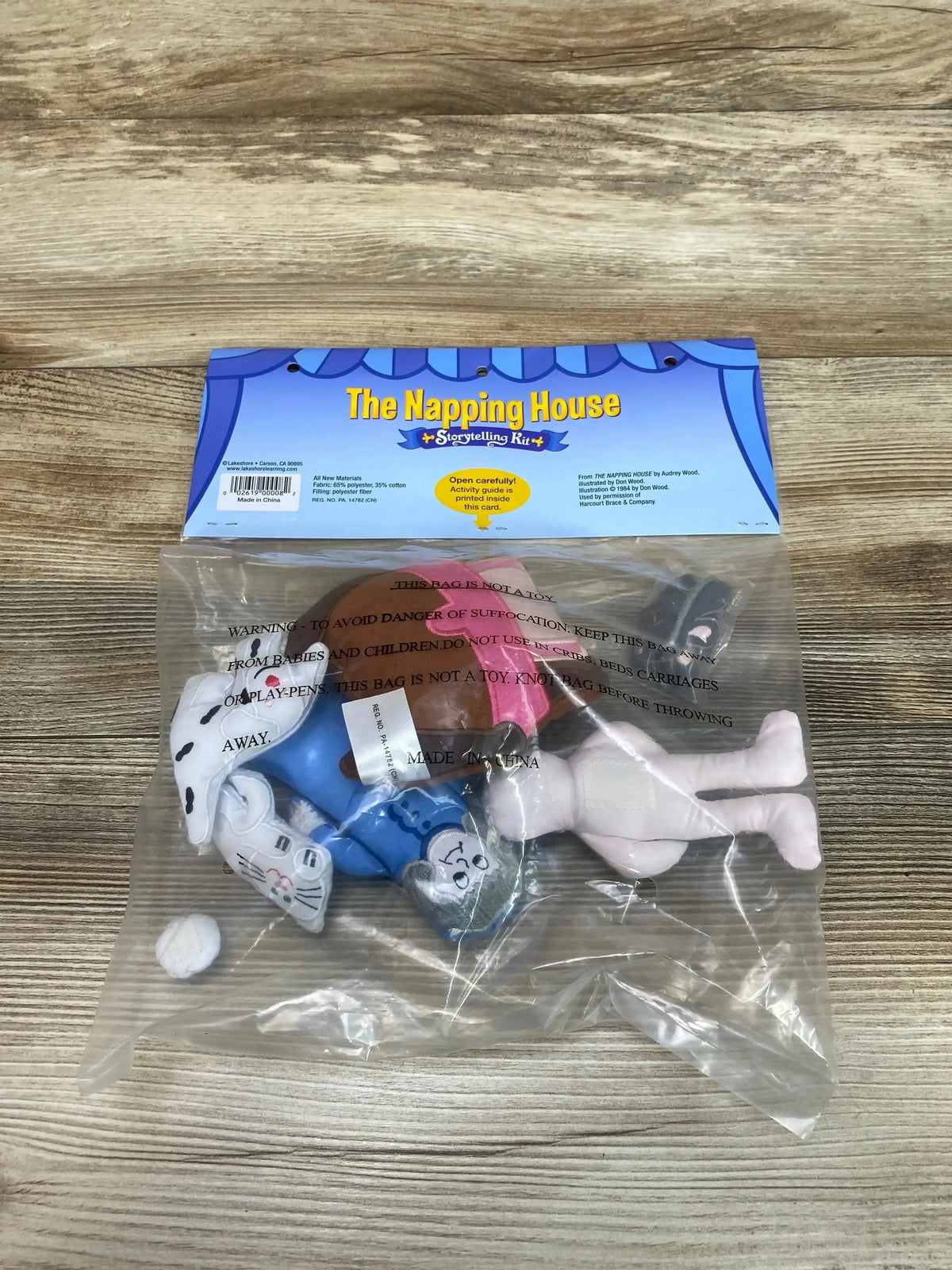 Top-down view of Lakeshore The Napping House Storytelling Kit with soft character puppets in clear bag on wood surface