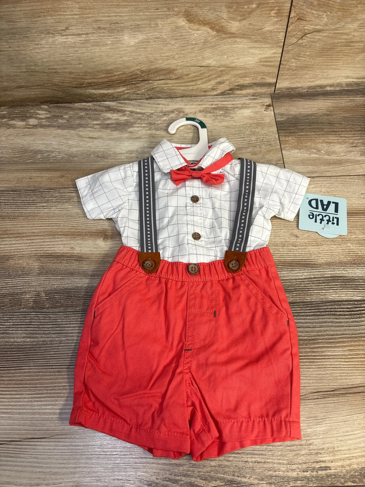Front view of Little Lad 4-piece suspender set showing checked bodysuit, red shorts, bowtie, and suspenders on a wood floor