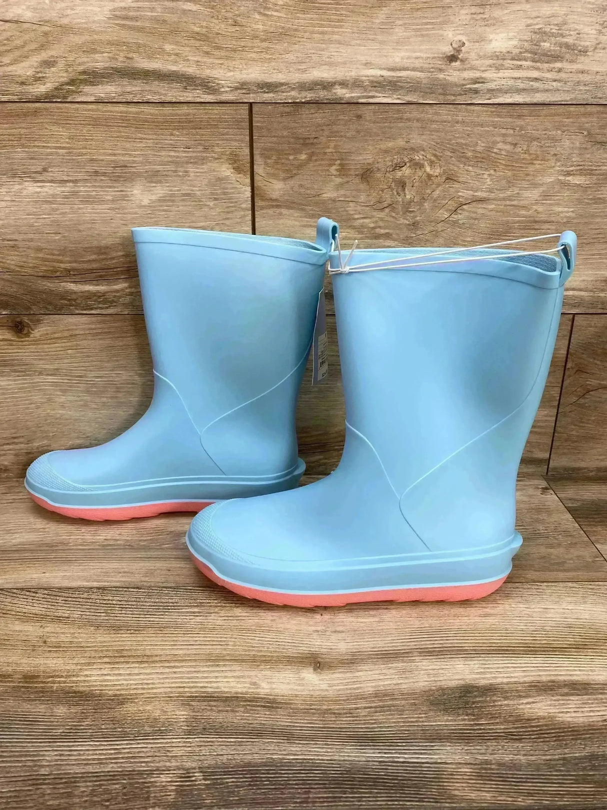 Side view of Cat and Jack Andy kids light blue slip-on rain boots size 6Y showcasing coral-colored soles and pull tabs on wood background