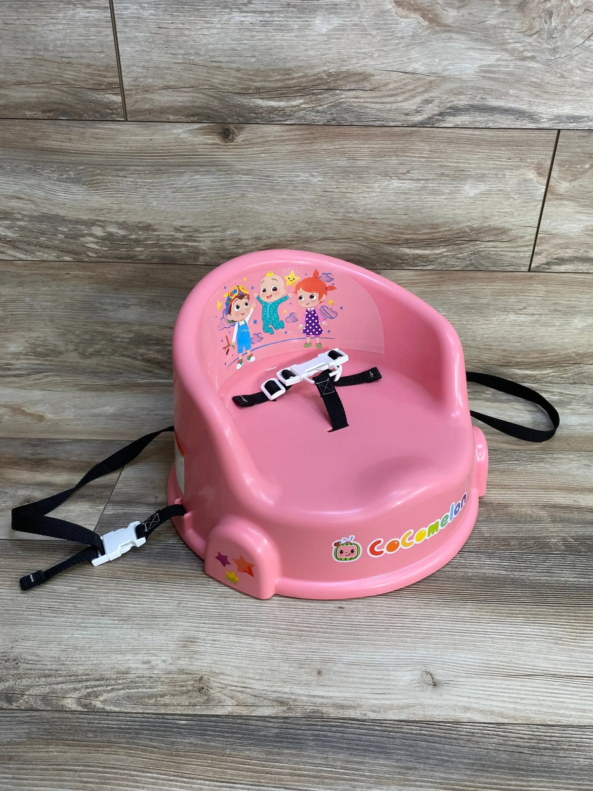 Three-quarter view of pink Cocomelon 15-inch booster seat with safety harness and character decals on wood background