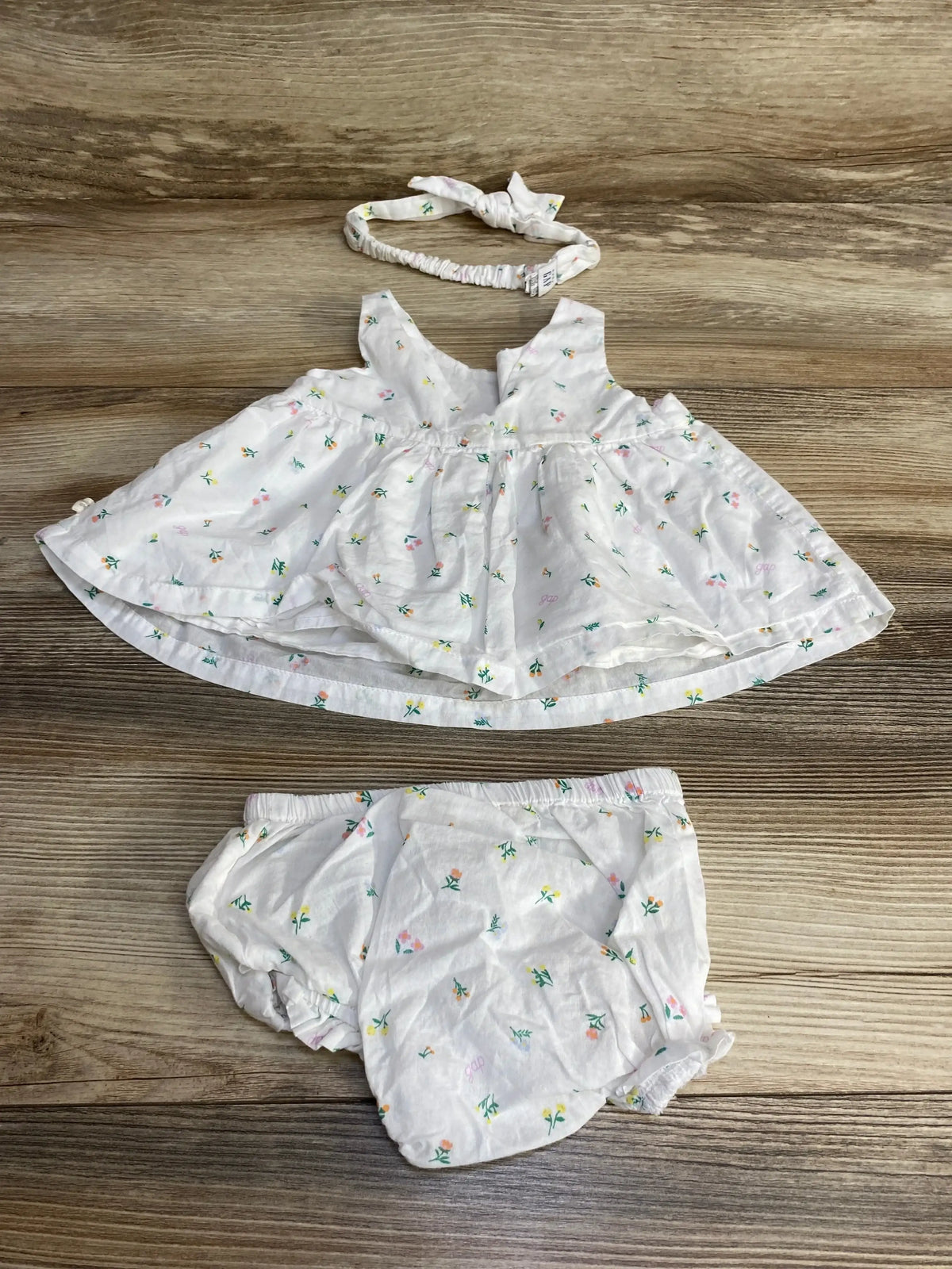 Flat lay of 3-piece Baby Gap white floral dress set showing gathered-waist sleeveless dress, elastic bloomers, knotted headband on wood background