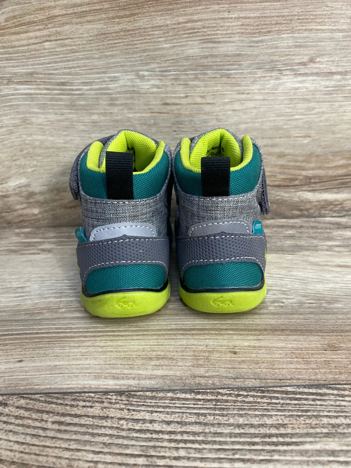 Rear view of See Kai Run Sam II waterproof boots, grey and teal with neon yellow soles and Velcro straps on wood background
