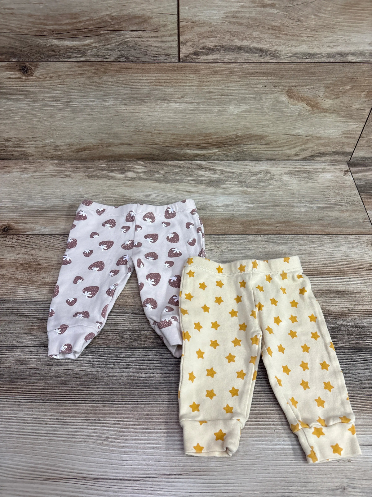 Top view of two baby pants in a 0 to 3 month set, one with yellow stars and one with brown strawberry print on wood flooring