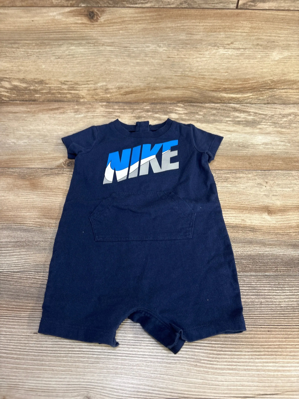 Front flat lay of navy Nike shortie romper (6 months) showing large NIKE logo and front kangaroo pocket on wood background