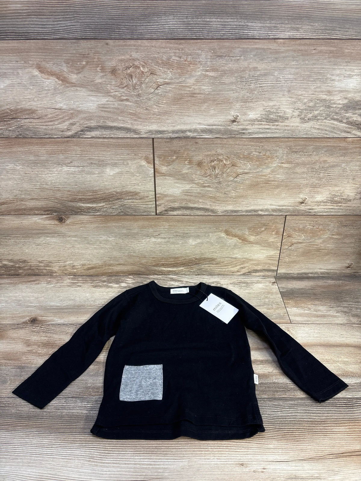 Front view of black Miles Baby long sleeve shirt showcasing a gray front pocket on a wood plank background