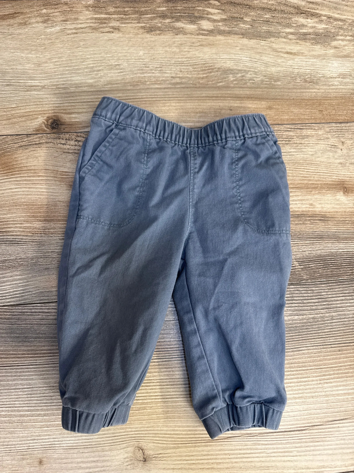 Flat lay of blue infant pull-on pants, size 3-6 months, showing elastic waistband and cuffed ankles on wood background